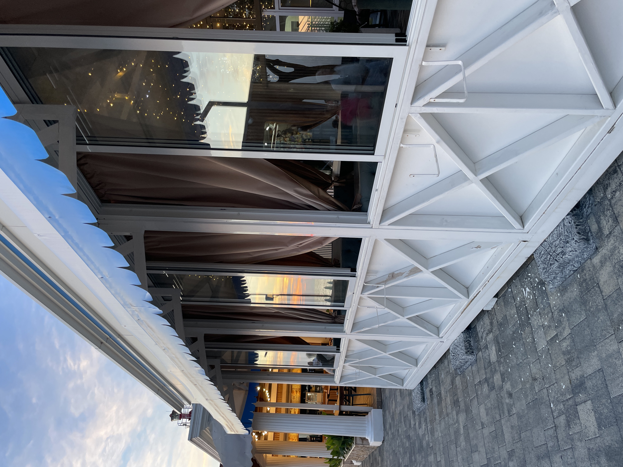 The image shows a white-paneled building with large windows and a blue awning, located in Sochi, Russia. The building appears to be a cafe or restaurant by the sea. The windows reflect the sky and the sea at sunset, with soft clouds and warm colors. Inside, the reflections show string lights and brown curtains. The building is situated on a paved walkway, and there are decorative white latticework panels on the lower section of the building. In the background, the facade of another building with white columns is visible. Several gray stone blocks are placed under the base of the white-paneled structure. There is no visible activity or people in the immediate foreground or on the walkway.