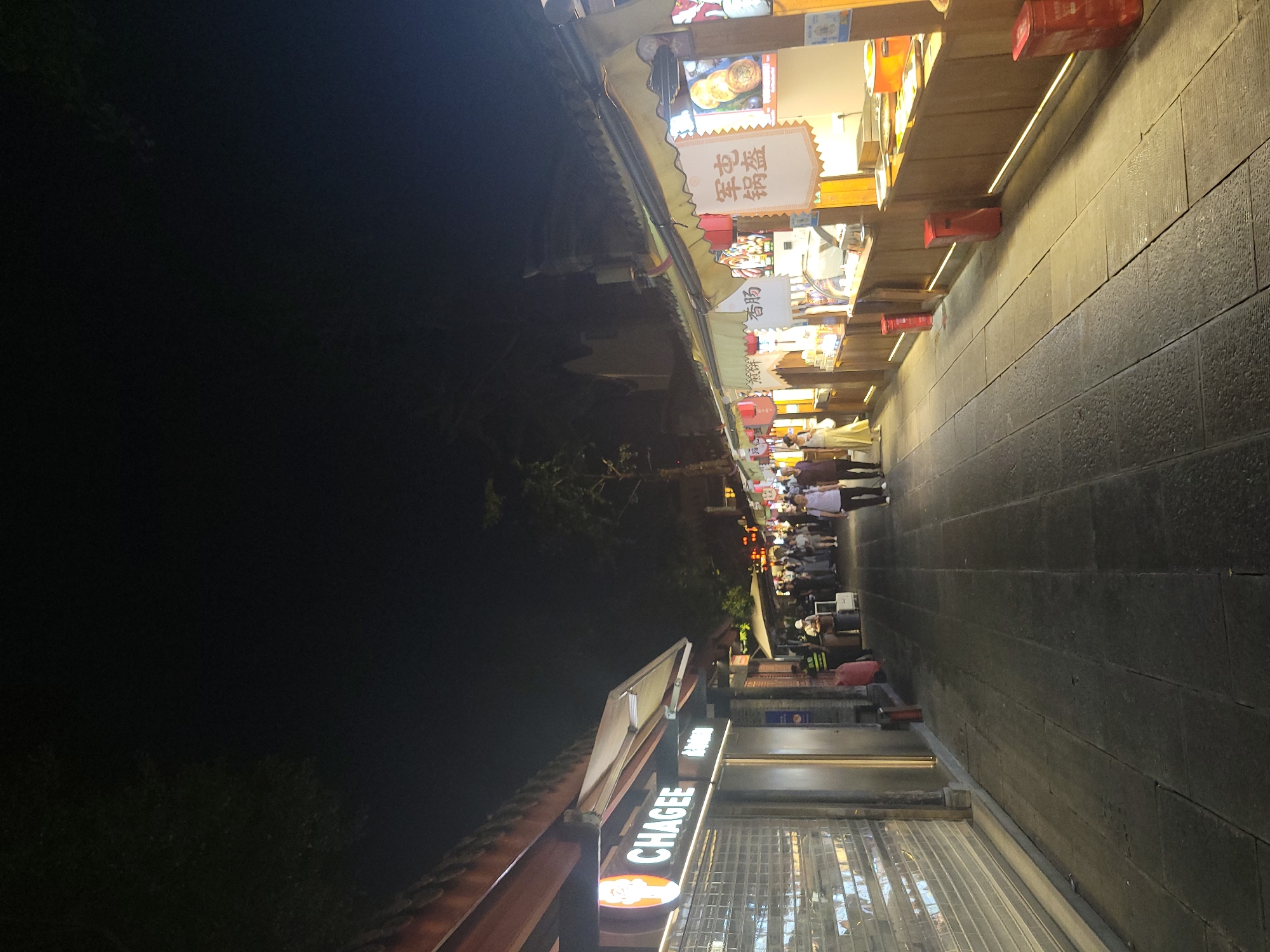 This is a nighttime, eye-level, full-shot photograph of a bustling pedestrian street lined with food stalls and shops in Wuhou District, China. The street is paved with dark, rectangular stones, and illuminated by the warm glow of lights from the stalls and street lamps. The sky above is dark, with a hint of tree foliage visible in the upper left corner.
On the left side of the street, a store with a prominent "CHAGEE" sign is visible. Its entrance is covered by a metallic roll-down gate. Further down the street, several figures are present, including a security guard wearing a reflective vest, and a few pedestrians browsing at the stalls.
The right side of the street is dominated by a row of food stalls. These stalls have wooden counters and are adorned with various signs, some featuring Chinese characters and images of food items. Red lanterns hang from some of the stall awnings. People are seen milling around the stalls, presumably purchasing or consuming food. The stalls are brightly lit, showcasing food displays and cooking equipment. Red cylindrical trash cans are placed along the sidewalk in front of some of the stalls. The overall atmosphere is lively and vibrant, characteristic of a busy night market.