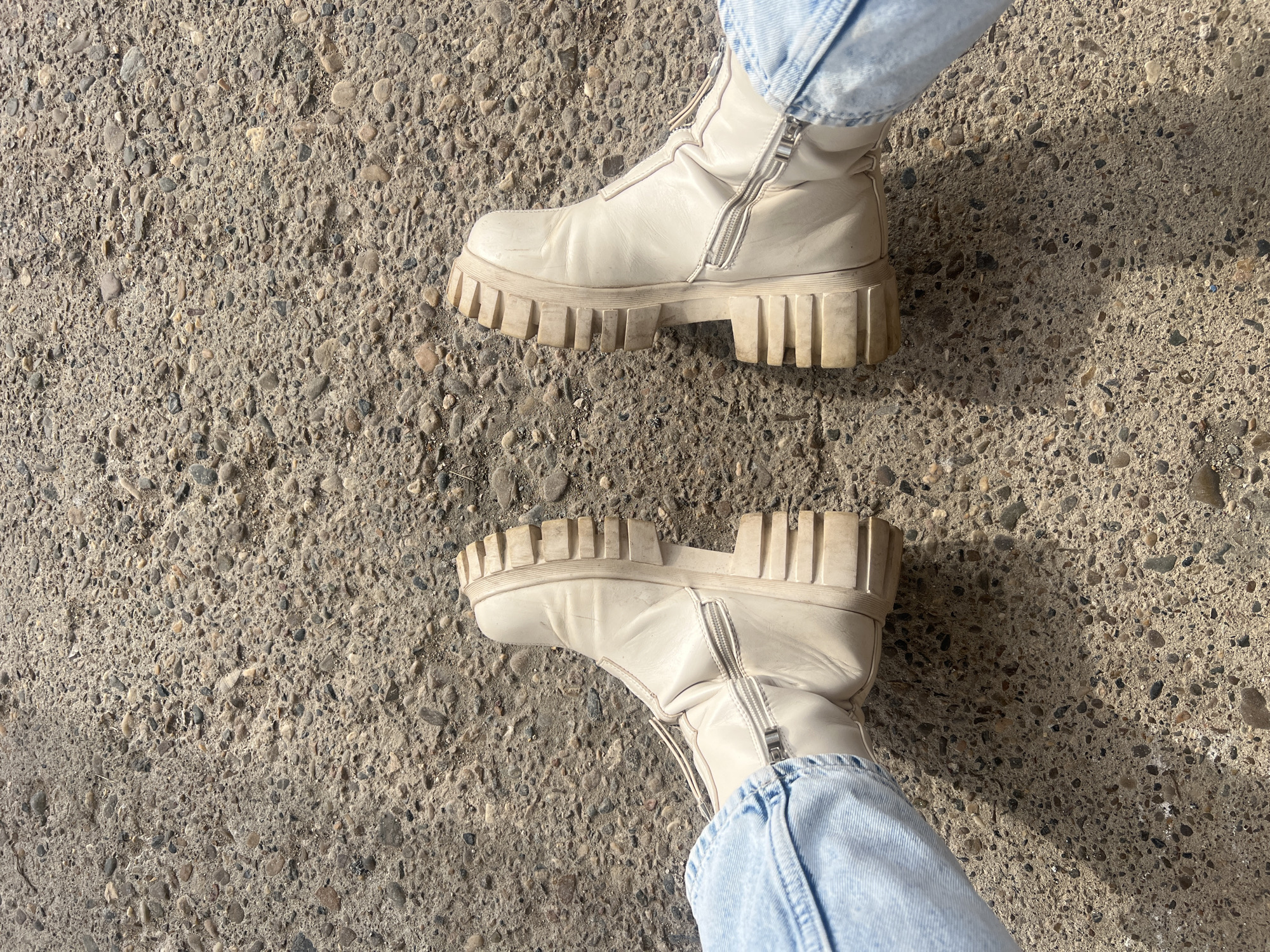 The image depicts a first-person perspective view of a person's lower legs and feet, presumably in Chaikovsky, Russia. The individual is wearing light blue denim jeans and a pair of cream-colored, ankle-high boots with substantial, chunky soles featuring deep horizontal treads. Each boot has a prominent silver-toned zipper on the inner side. The feet are positioned slightly apart, with the toes angled generally upwards. The underlying surface is a textured, light grey concrete or asphalt path, embedded with numerous small, multi-colored pebbles and aggregate. Patches of sunlight and shadow are visible on the ground, indicating an outdoor setting during daylight hours. No other individuals or significant objects are present within the frame.