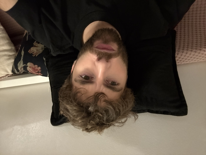 A bearded man with curly hair is lying on a bed or couch, his head resting on a black pillow. He is looking directly at the camera with a neutral expression. He is wearing a black shirt. Two decorative pillows are visible in the background: one is a patterned floral pillow and the other is a light pink textured pillow. The wall behind the bed is white.