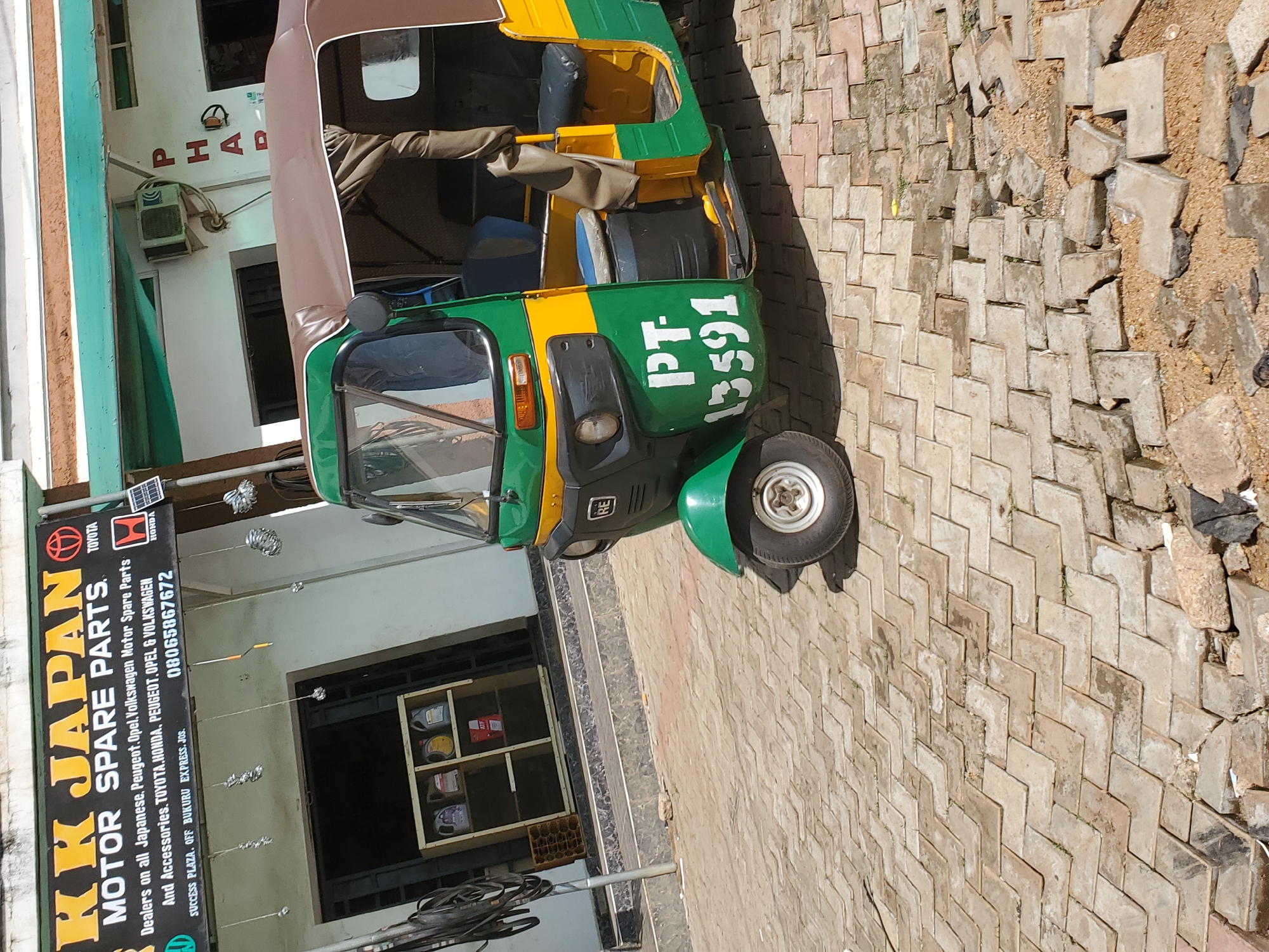 The image depicts the exterior of a business named "KK JAPAN MOTOR SPARE PARTS" located at Success Plaza, off Bukuru Express, Jos, Nigeria. A green and yellow three-wheeled auto-rickshaw, also known as a "Keke Napep" or "Tuk-tuk," is parked in front of the shop. The auto-rickshaw has the number "PT-13591" painted on its side. The shop sign displays various motor brands like Toyota and Honda. The pavement in front of the shop is paved with interlocking concrete blocks, some of which are damaged or missing, revealing sand and debris underneath. The overall scene suggests a commercial area in Nigeria, possibly a street in Bukuru, Jos.