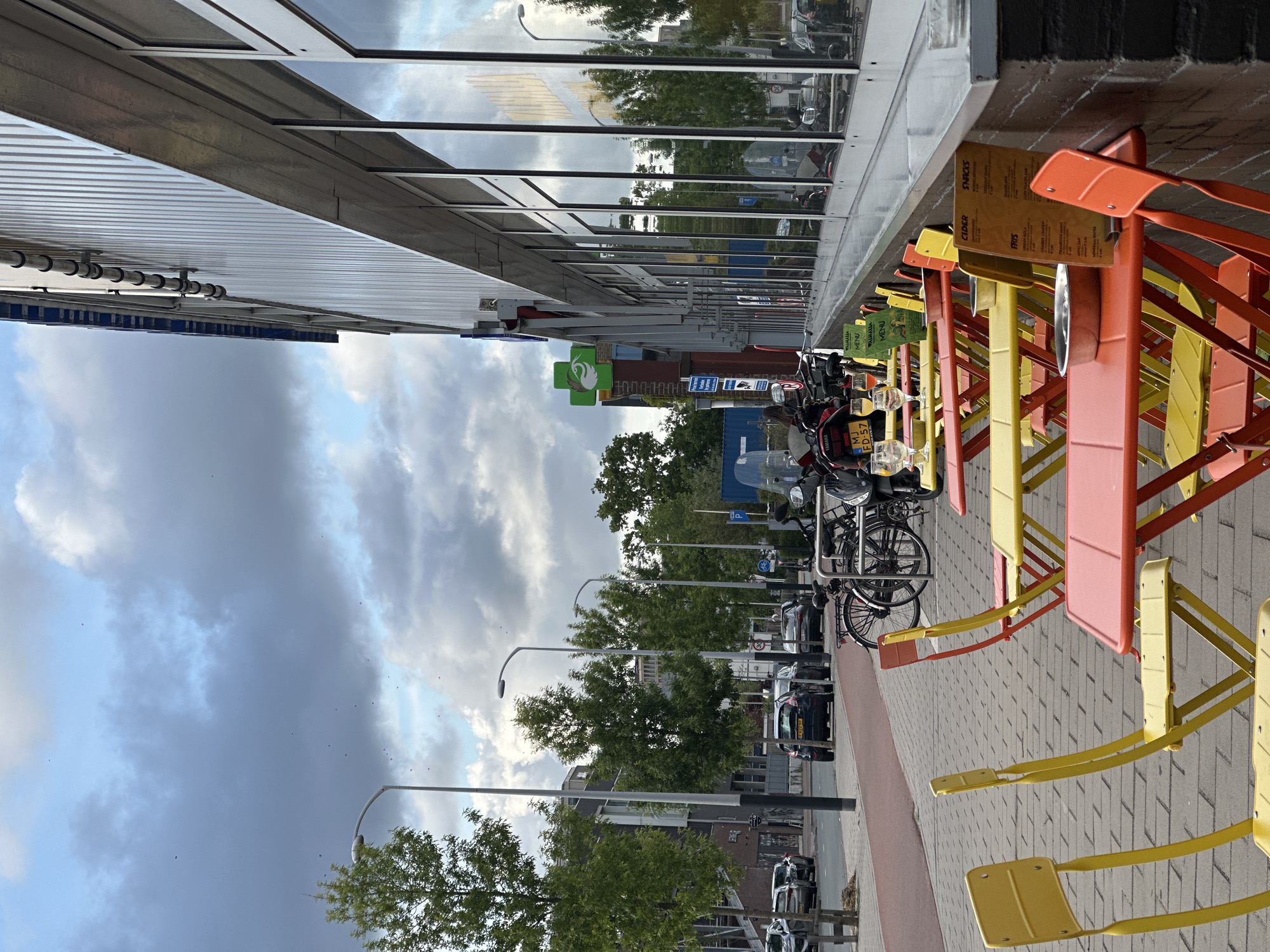 The image depicts an outdoor scene in Amsterdam, Netherlands, along a street lined with buildings, trees, and parked vehicles. On the right side of the frame, several colorful tables and chairs (yellow, orange, and red) are arranged on the sidewalk, seemingly part of a cafe or restaurant's outdoor seating area. Menus are attached to the chairs. Bicycles and motorbikes are parked nearby. Across the street, there's a row of parked cars visible. Street lamps are also present along the street. The sky above is overcast, with dark clouds. The overall atmosphere suggests an urban setting on a potentially cloudy day. A green pharmacy sign is visible in the background. The scene is captured from a perspective close to the ground, with an upright orientation.