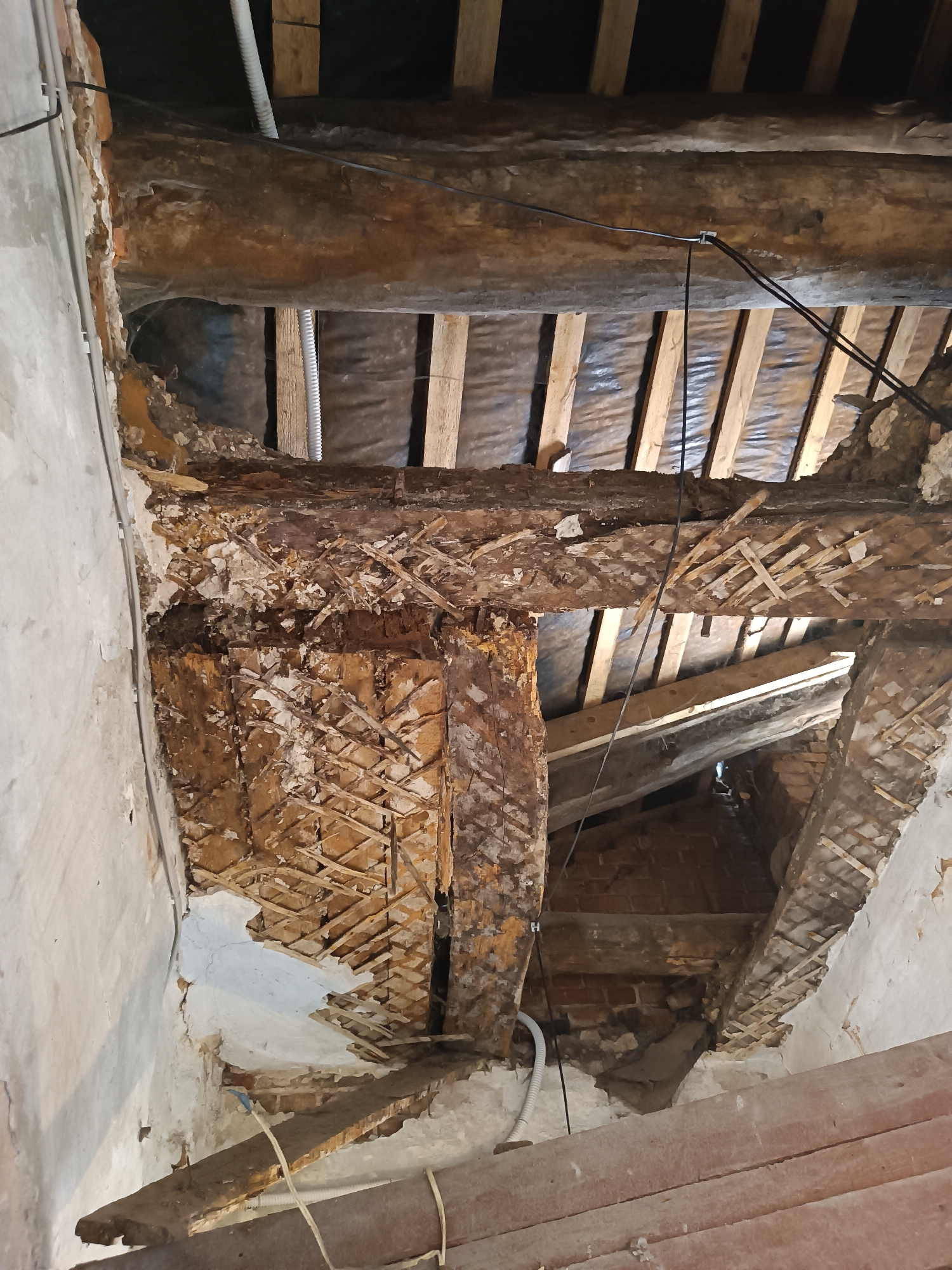 The image depicts the interior of a building undergoing renovation or repair, likely in the city of Yelets, Russia. The focus is on damaged wooden beams and supporting structures that appear to be part of a ceiling or roof assembly. Several large, aged wooden beams are visible, some showing signs of rot, decay, and structural compromise. Exposed lath and plaster remnants suggest that a plaster ceiling was once attached to these beams.
In the background, parallel wooden rafters are visible, covered with a dark material, possibly insulation or a vapor barrier. Electrical conduits and cables are strung between the beams, indicating ongoing or recent electrical work. The overall environment appears dusty and unfinished, with debris scattered around. There are no people present in the image. The scene suggests structural damage and a process of demolition or reconstruction of the building's internal framework.