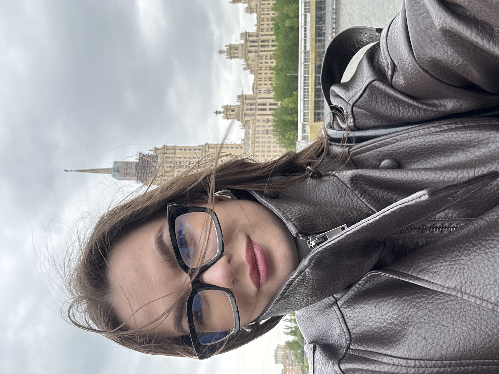 A woman wearing eyeglasses and a leather jacket is pictured in front of the Moscow State University building in Moscow, Russia. The background features the main building of Moscow State University, characterized by its Stalinist architecture and prominent spire. The weather appears overcast.