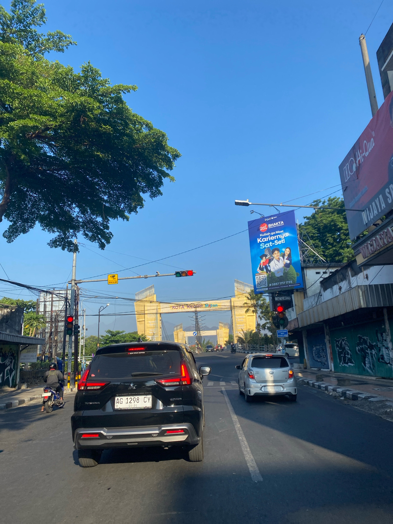The image captures a street scene in Kediri City, Indonesia, under a clear blue sky. A black SUV, with license plate AG 1298 CY, is in the foreground, facing away from the camera. To its right, a silver car is also traveling away. On the left side of the road, a person on a motorcycle is visible. Overhead, traffic lights are present, with the red light illuminated. In the background, a large yellow archway structure spans the road, bearing the text "KOTA KEDIRI". A billboard for "BHAKTA WIJAYA" is also prominently displayed. The street is lined with buildings and trees, and power lines crisscross the scene. The overall impression is of a typical urban environment during daylight hours.