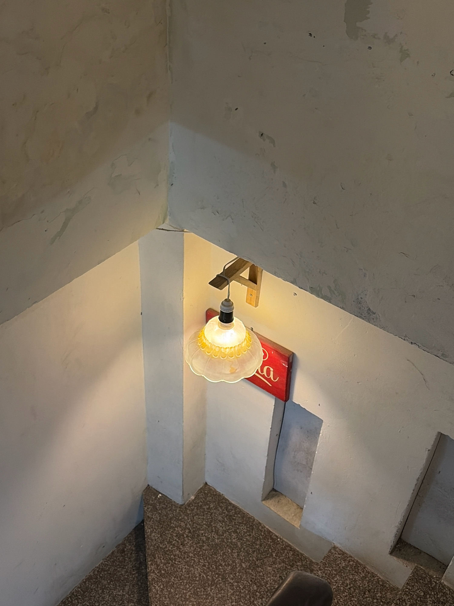 The image shows a dimly lit staircase with speckled brown carpeted steps. A vintage-style light fixture with a clear glass shade hangs from the ceiling, casting a warm glow. Below the light, a red sign with white script is mounted on the wall. The walls are textured and appear to be painted a pale color. The scene suggests an interior space, possibly a cafe or residential building. While the specific location in Da Nang, Vietnam is not explicitly indicated, the overall aesthetic hints at a potentially older or characterful establishment.