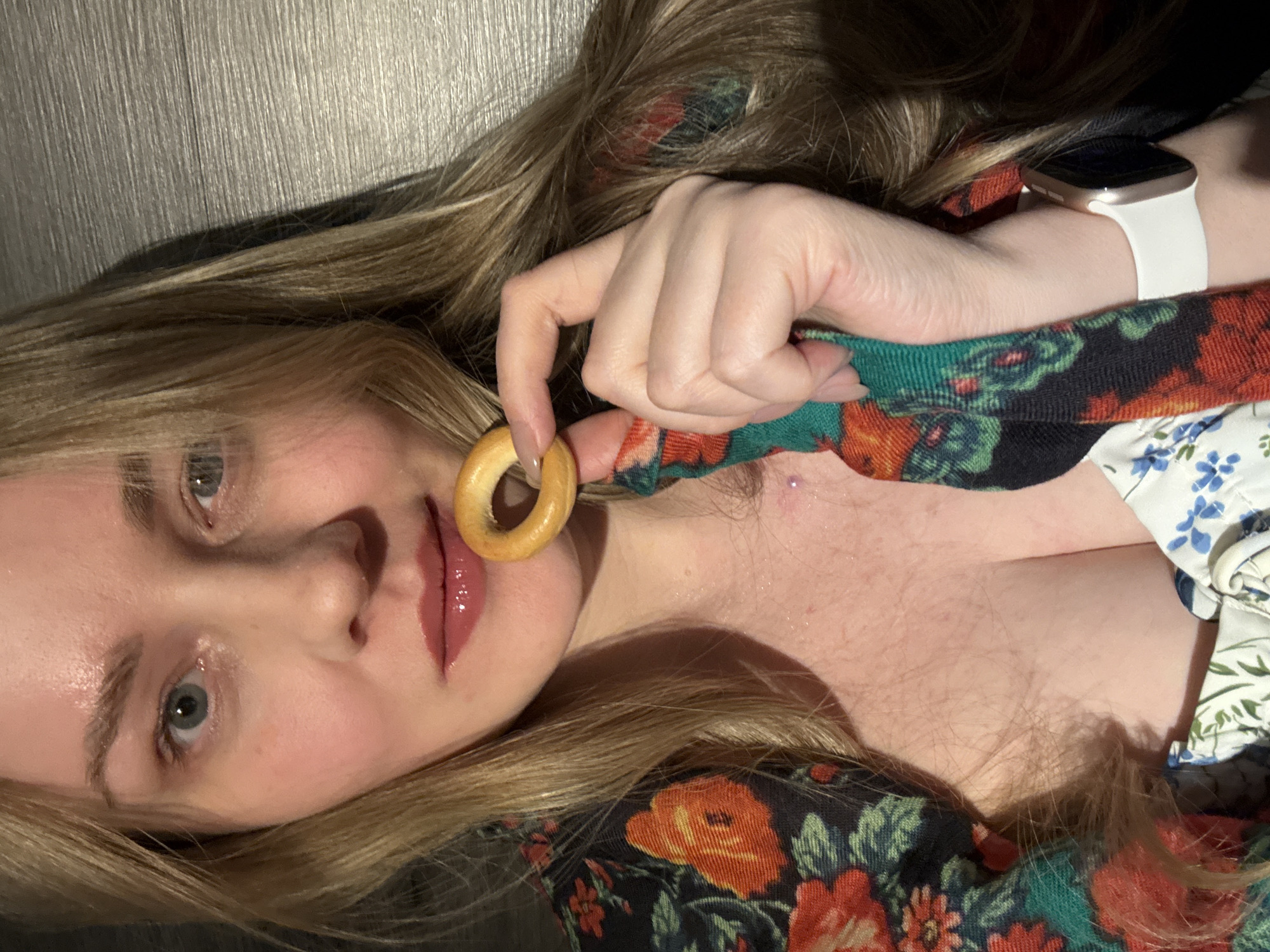 A woman with fair skin and blonde hair is holding a small, circular bagel near her lips. She is wearing a floral print dress and a white smartwatch on her left wrist. The background appears to be a dark, flat surface. There is no information to indicate a location in the city of Chaikovsky, Russia.