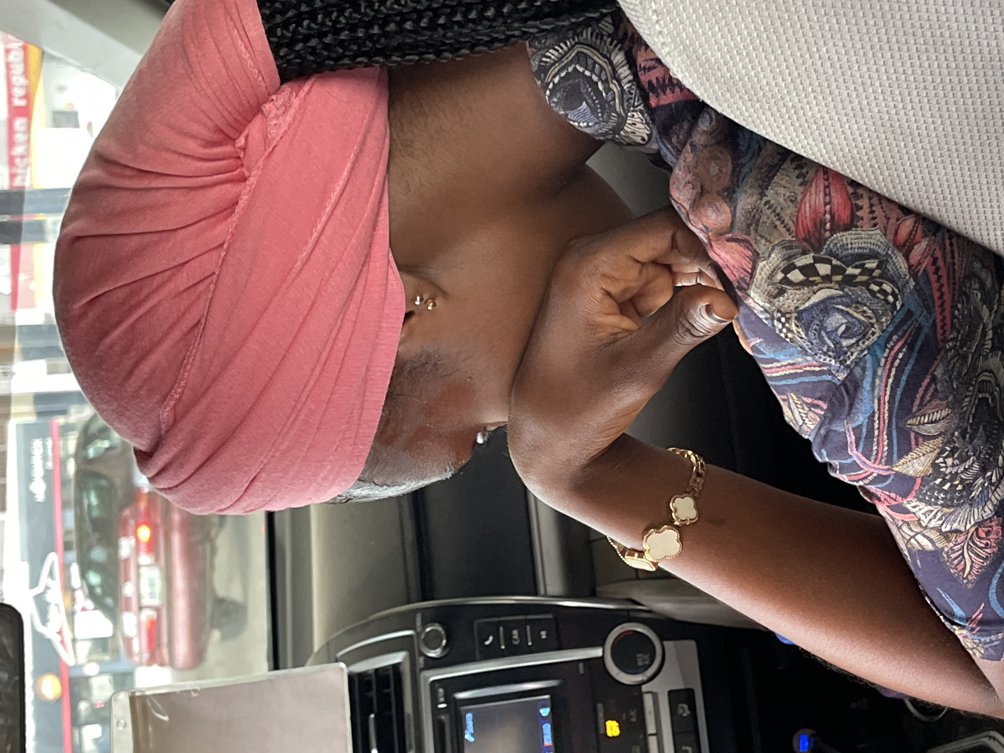 This is a close-up, vertically oriented photo of a woman wearing a pink headscarf and a patterned shirt. She is seated in what appears to be the passenger seat of a car, with a portion of the car's dashboard visible in the lower part of the frame.
The woman's right arm rests on her face, and she is wearing a gold bracelet with white clover-shaped charms. She has a subtle smile, suggesting a relaxed or content mood. The lighting indicates it might be daytime, possibly afternoon, given the natural light entering the car.
Behind the woman, reflections in the car window show what appears to be a street scene with some traffic, including a red vehicle. The pattern on her shirt is colorful and intricate, featuring shades of blue, pink, red, and yellow.
The scene is set inside a car, likely in an urban or semi-urban environment based on the reflections. There are no explicit emotional expressions captured, beyond the subtle smile. The time of day is indeterminate, but the lighting suggests it is not nighttime. The location cues are limited to the interior of a car and the street scene outside.