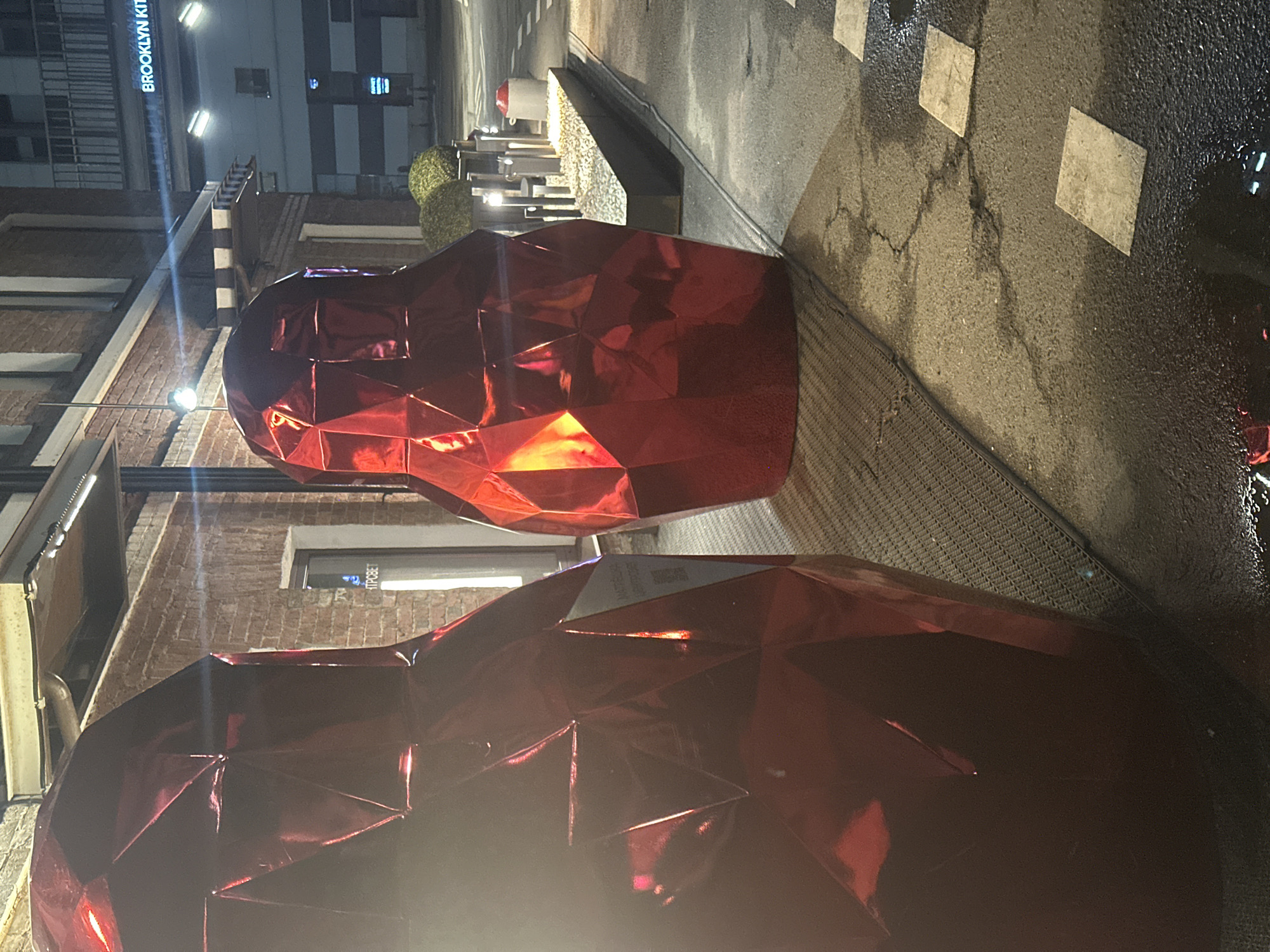 The image depicts a nighttime scene featuring two large, shiny red polygonal sculptures, possibly hearts, located on a sidewalk in Moscow, Russia. The architecture visible in the background suggests an urban environment, with buildings and streetlights present. A sign reading "Brooklyn Kitchen" is seen vertically on a building in the background. The sidewalk is patterned with squares, possibly indicating a pedestrian crossing. The lighting in the scene creates reflections on the sculptures and wet areas of the sidewalk.