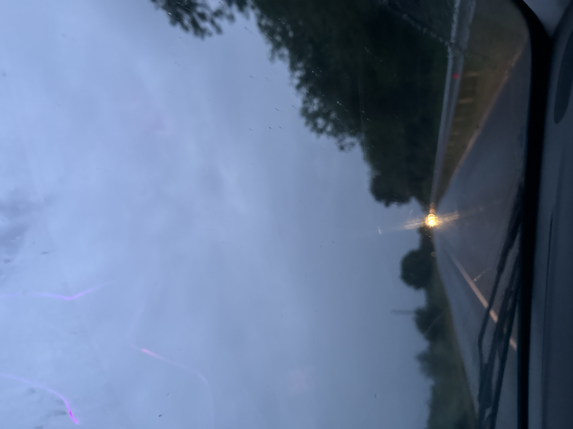 The image is taken from inside a vehicle, looking out through the windshield. It appears to be a cloudy or overcast day. The scene depicts a road ahead, with trees bordering the side of the road. The road is wet. There's a headlight shining at the center of the road in the distance. The wipers are visible in the bottom right. There is no information to suggest that the image was taken in Zvanichevo, Bulgaria.