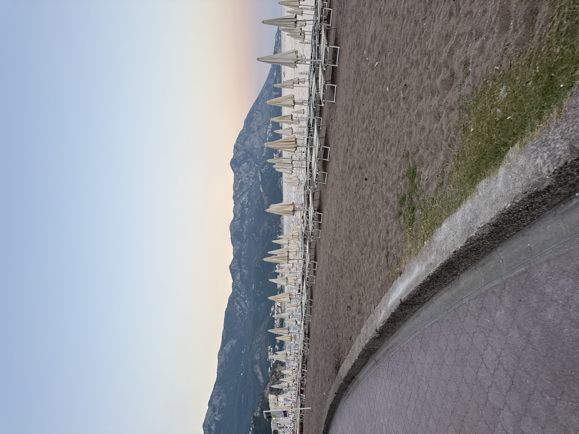 The image shows a view of the beach in Boreti, Montenegro, at dusk. The sand is dark and appears to be volcanic. Rows of closed beach umbrellas and lounge chairs are neatly arranged on the sand, facing the calm, dark water of the Adriatic Sea. In the background, a range of green, tree-covered mountains rises above the sea, with a few buildings visible on the lower slopes. The sky is a soft gradient of pale blue and a hint of pink, indicating the transition from day to night. A paved walkway with a textured surface and a concrete border curves along the left side of the frame, separating the beach from the foreground. No people are visible in the scene.