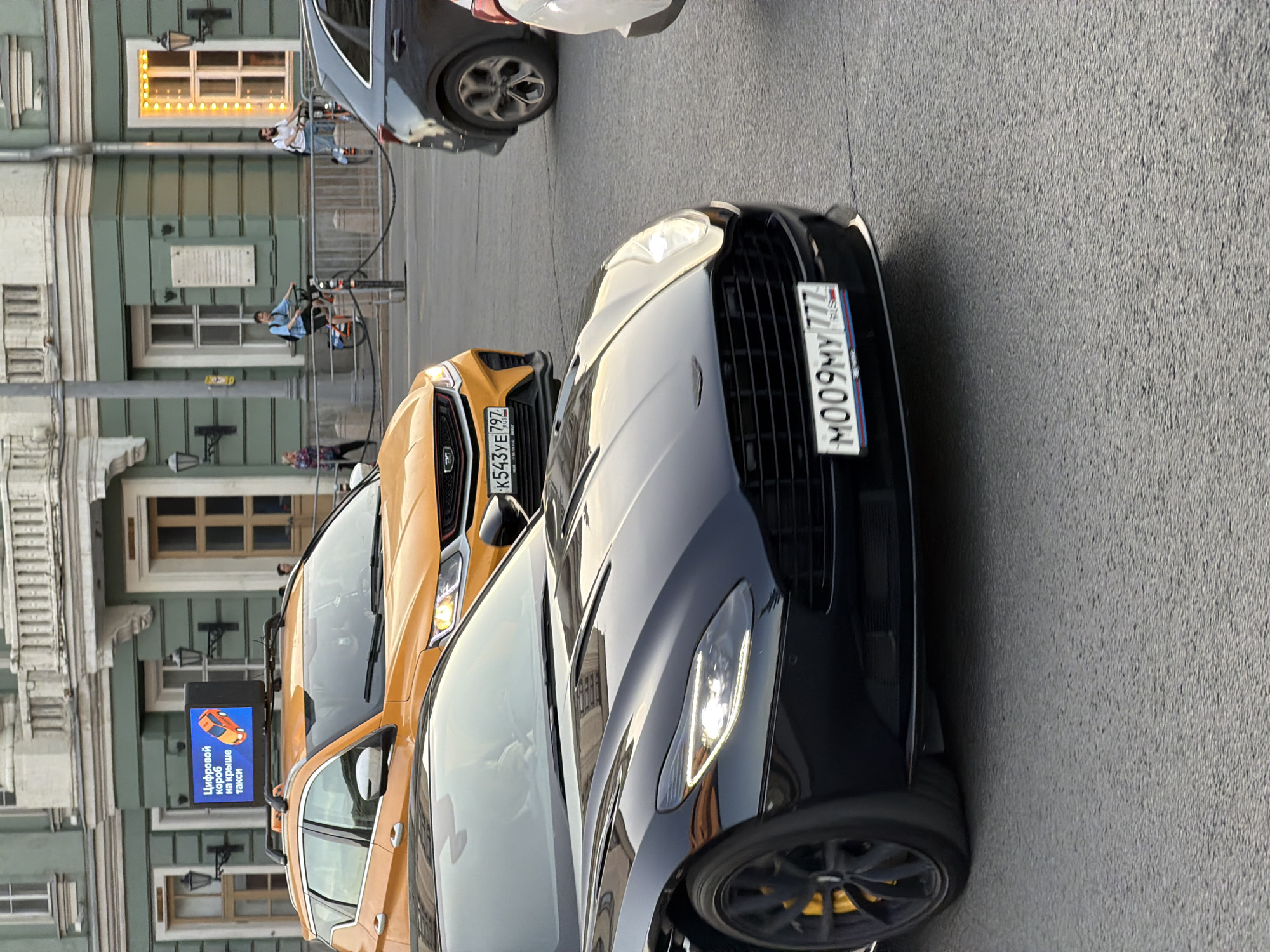 The image captures a street scene in Moscow, Russia. A black Aston Martin sports car, bearing the license plate "M009MY 777," is prominently positioned, parked adjacent to an orange Geely Tugella with license plate "K543YE 797." A dark SUV is partially visible behind the Aston Martin. The vehicles are parked along a street lined with buildings constructed in a classical architectural style, painted in shades of green and beige. A sign indicating a taxi service with the text "Цифровой короб на крыше такси" (Digital box on the roof of a taxi) is attached to the Geely Tugella, suggesting its use as a taxi. Several pedestrians are seen on the sidewalk in the background, and bicycles secured by a fence along the sidewalk. Overhead streetlights and ornate architectural details on the buildings further define the scene as an urban setting.