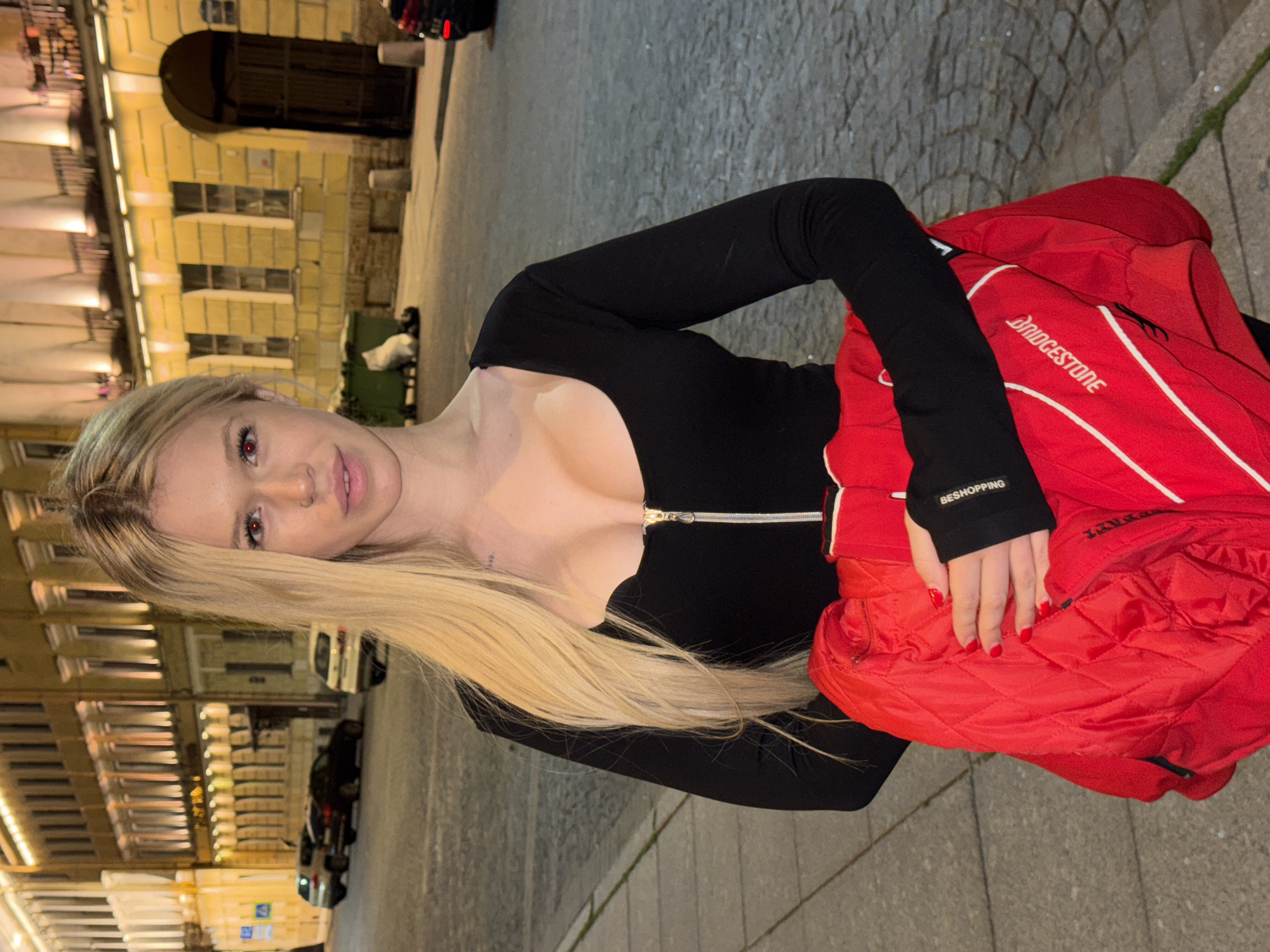 A young woman with blonde hair and red eyes stands on a cobblestone sidewalk at night. She is wearing a black, long-sleeved top with a zipper and a bright red quilted jacket draped over her arm. The jacket has white and red stripes and white lettering that reads "BRIDGESTONE". In the background, a street lined with buildings illuminated by warm lights stretches into the distance. Cars are parked along the street, and green trash bins are visible. The architecture suggests the scene is in Saint Petersburg, Russia, likely near the historic center given the building style and cobblestone streets.