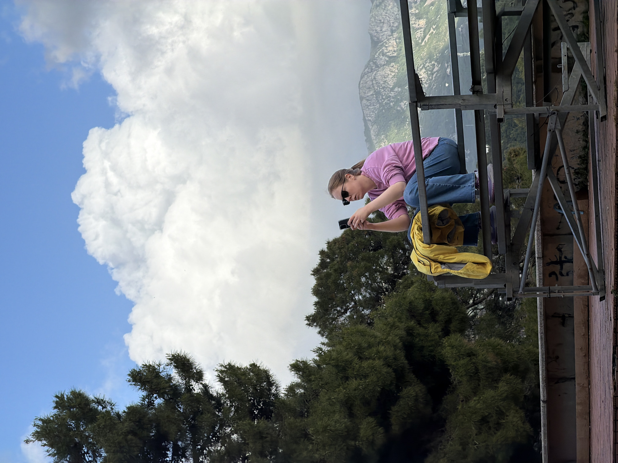 A woman with light hair pulled back in a ponytail, wearing sunglasses, a pink long-sleeved shirt, and blue jeans, is crouching on a metal structure, possibly scaffolding or a platform, while looking at and holding a cell phone in her hands. A yellow jacket is draped on a portion of the metal structure next to her. The metal structure appears to be affixed to a brick or concrete wall with graffiti on it. In the background, there are green trees and a partially cloudy sky. In the distance, there is a mountain range. Based on the landscape and architecture visible, it is likely this photo was taken in the city of Budva, Montenegro.