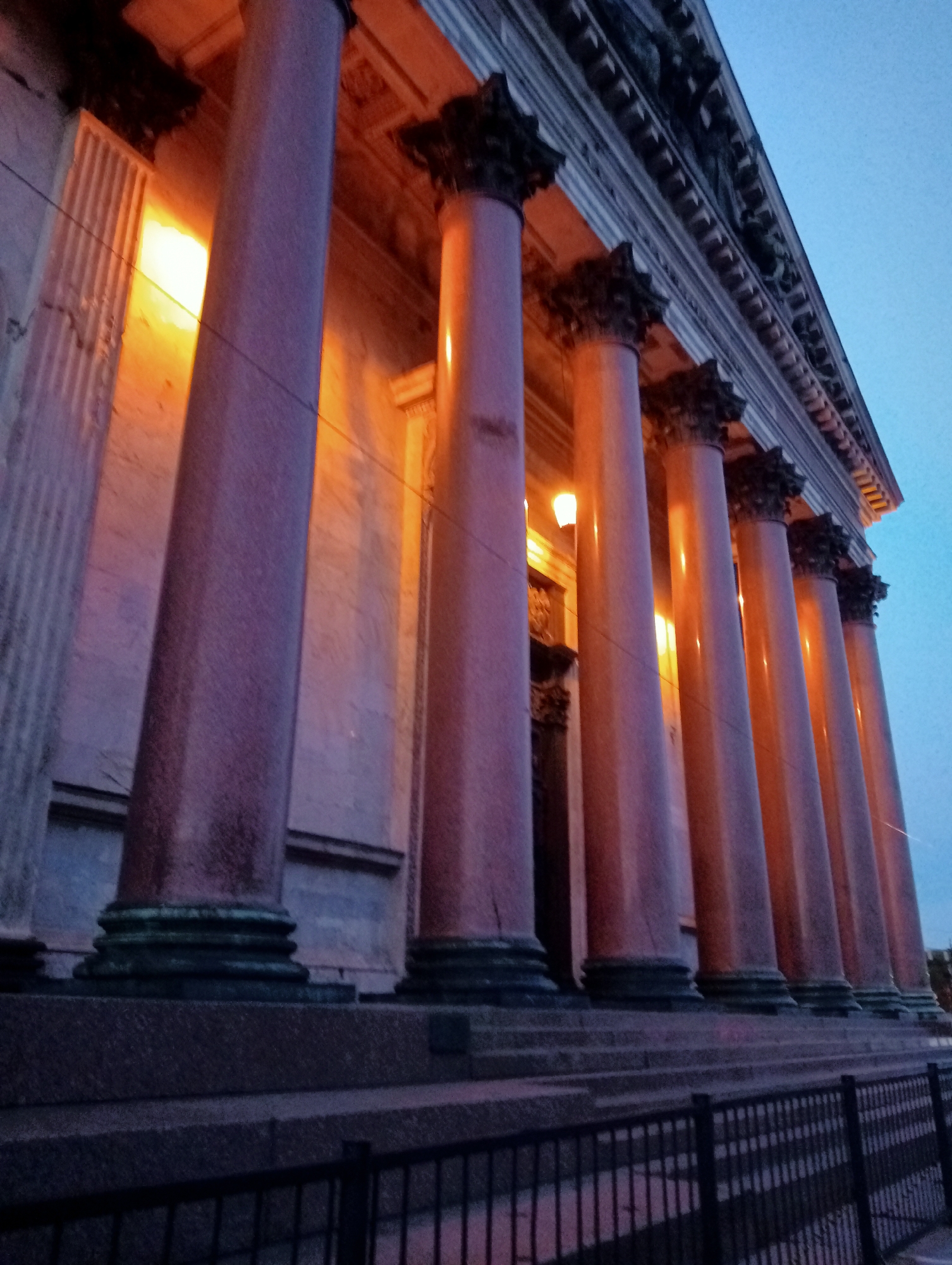 The image shows a low-angle, wide shot of the exterior of a grand neoclassical building, likely St. Isaac's Cathedral in Saint Petersburg, Russia, at dusk. The building features a colonnade of several imposing pink granite columns with dark, ornate capitals. Warm light emanates from within the building, illuminating the marble facade and casting a glow on the columns. The sky above is a deep twilight blue. In the foreground, there is a dark metal fence, and a stone staircase leads up to the building's entrance, partially obscured by the columns. The scene conveys a sense of historical grandeur and architectural majesty. There are no people visible in the image.