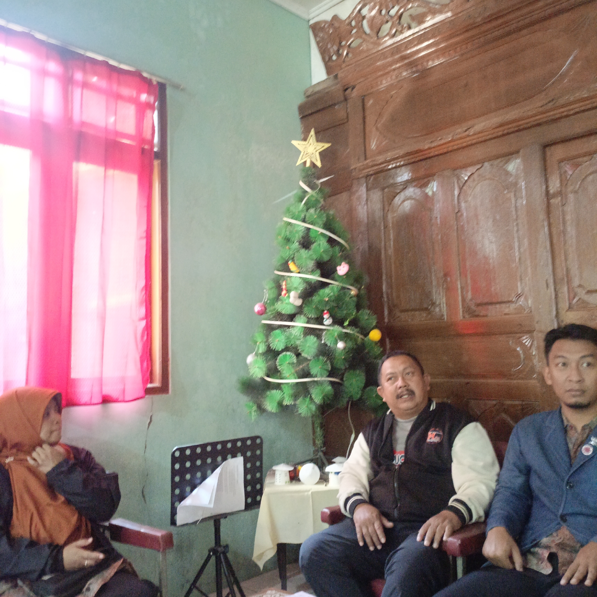 In an indoor setting, likely a living room in Salatiga, Indonesia, a small, artificial Christmas tree decorated with white ribbon and various ornaments stands prominently. A golden star adorns the top of the tree. To the left, a woman wearing a brown hijab and an orange headscarf is seated, her hand near her face, appearing to be adjusting her headscarf or touching her cheek. In front of her is a black music stand with a white paper sheet. Three men are seated to the right of the tree. The man closest to the tree is wearing a black and white jacket over a grey shirt, and dark pants, with a mustache. The man next to him is wearing a blue jacket over a patterned shirt and dark pants. Both men are looking towards the right of the frame, seemingly engaged in conversation or observing something outside the camera's view. A wooden cupboard with intricate carvings is visible behind the men and the Christmas tree. A window with pink curtains is to the far left, partially obscuring the view of what is outside. A small table covered with a cream-colored cloth is positioned behind the Christmas tree, holding some small decorative items. The wall color appears to be a pale green.