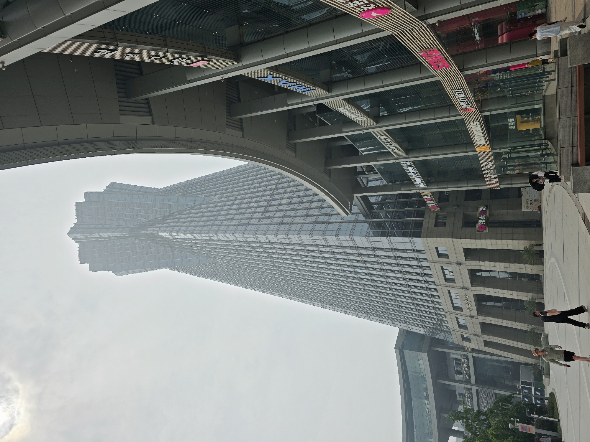 The image depicts the **Zhongtian Pavilion** in Gongshu District, China, on a hazy day. A very tall, modern skyscraper with a distinctive twisted design dominates the central and left portions of the frame. The skyscraper's facade is composed of glass panels and metallic elements.
In the foreground, a wide, paved plaza area is visible. To the right, a multi-story building with a curved, overhanging structure is present. This building features multiple levels of glass windows, with various signs indicating businesses such as "McDonald's," "luckin coffee," and "imax." The architectural style is modern and sleek.
Several people are present in the plaza. Two individuals are walking in the mid-ground, one wearing a hat and a light-colored jacket with dark shorts, the other in dark clothing. Another person is walking in the background to the right, wearing light-colored pants and a white top. Two individuals are visible near the entrance of the curved building, one holding an umbrella. Further in the background, a lone figure is seen walking.
The environment appears to be an urban setting with a focus on modern architecture. The sky is overcast, contributing to the hazy atmosphere. Greenery, including trees and plants, is visible to the left of the frame, adjacent to another building. The overall scene conveys a sense of a bustling commercial and urban center.