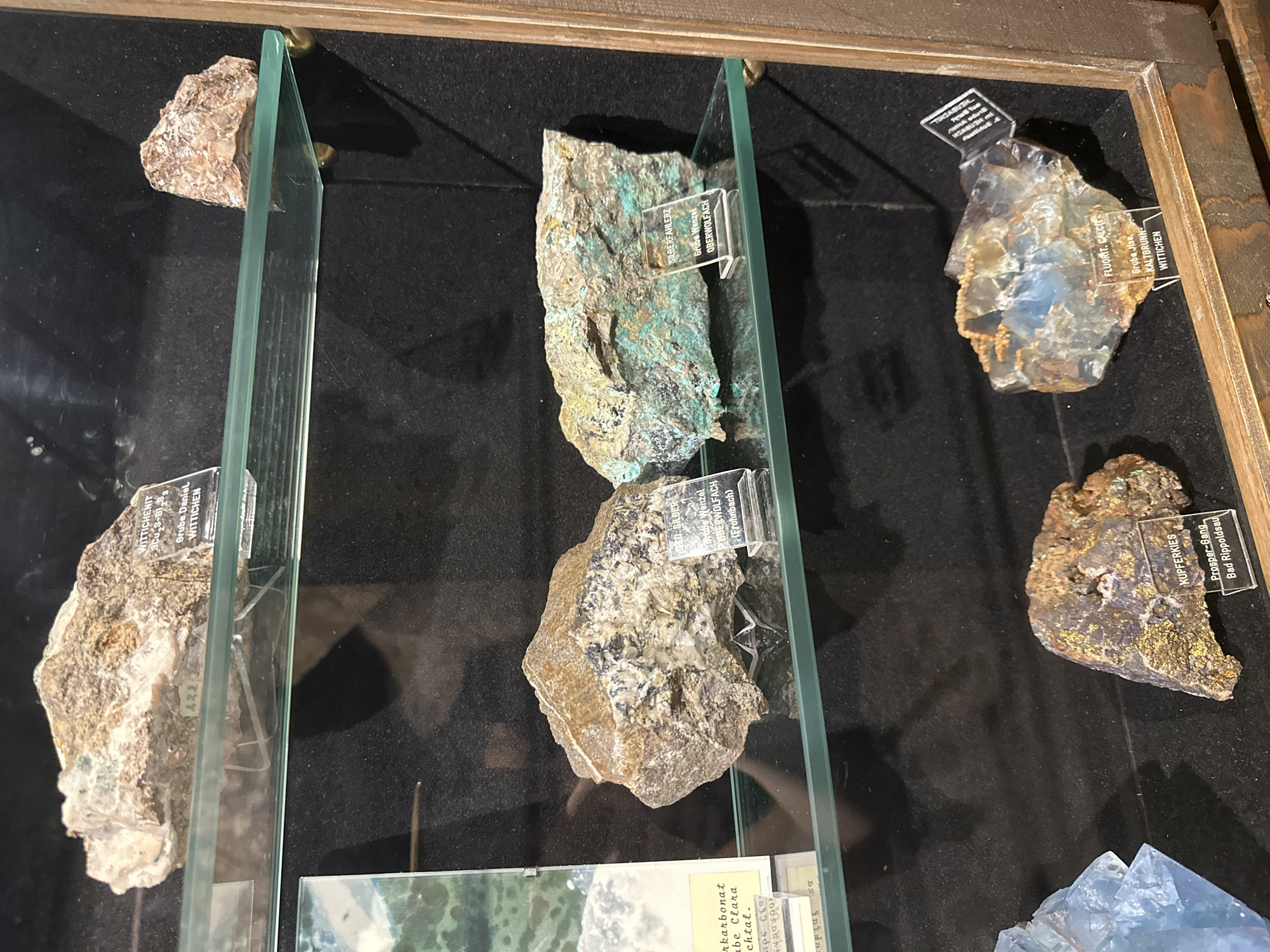 The image displays a collection of mineral specimens in a museum display case, likely located in Schonach im Schwarzwald, Germany, as indicated by labels on some of the specimens. The display case has glass shelves with several rocks and minerals.
On the top shelf, a rock labeled "WITTICHENIT Cu3 BiS3" is visible, along with another unlabeled rock on the right.
The middle shelf holds two larger specimens. The left specimen is a roughly textured rock with lighter and darker brown and gray tones. The right specimen is a large, irregular chunk of rock with prominent green and turquoise mineral deposits. Both have small, clear plastic labels. The left label is partially obscured but seems to read "GEIL SILBER" and "Grube Wenzel OBERWOLFACH (Frohnbach)". The right label clearly reads "SILBERFAHLERZ" and "Grube Wenzel OBERWOLFACH".
The bottom shelf features at least three specimens. On the left, a piece of blue, crystalline fluorite is visible. To its right, a brown, metallic-looking rock with flecks of gold is labeled "KUPFERKIES" and "Prosper-Gang Bad Rippoldsau". Further to the right, a translucent, multi-colored mineral specimen, possibly fluorite and calcite, is labeled "FLUORIT. CALCITE" and "Grube Ilse, KALBRUNN WITTICHEN". There's another unlabeled specimen to the far right, partially visible.
The background of the display case is dark, creating a contrast that highlights the mineral specimens. The overall presentation suggests a geological or mineralogical exhibition. No people are present in the image.