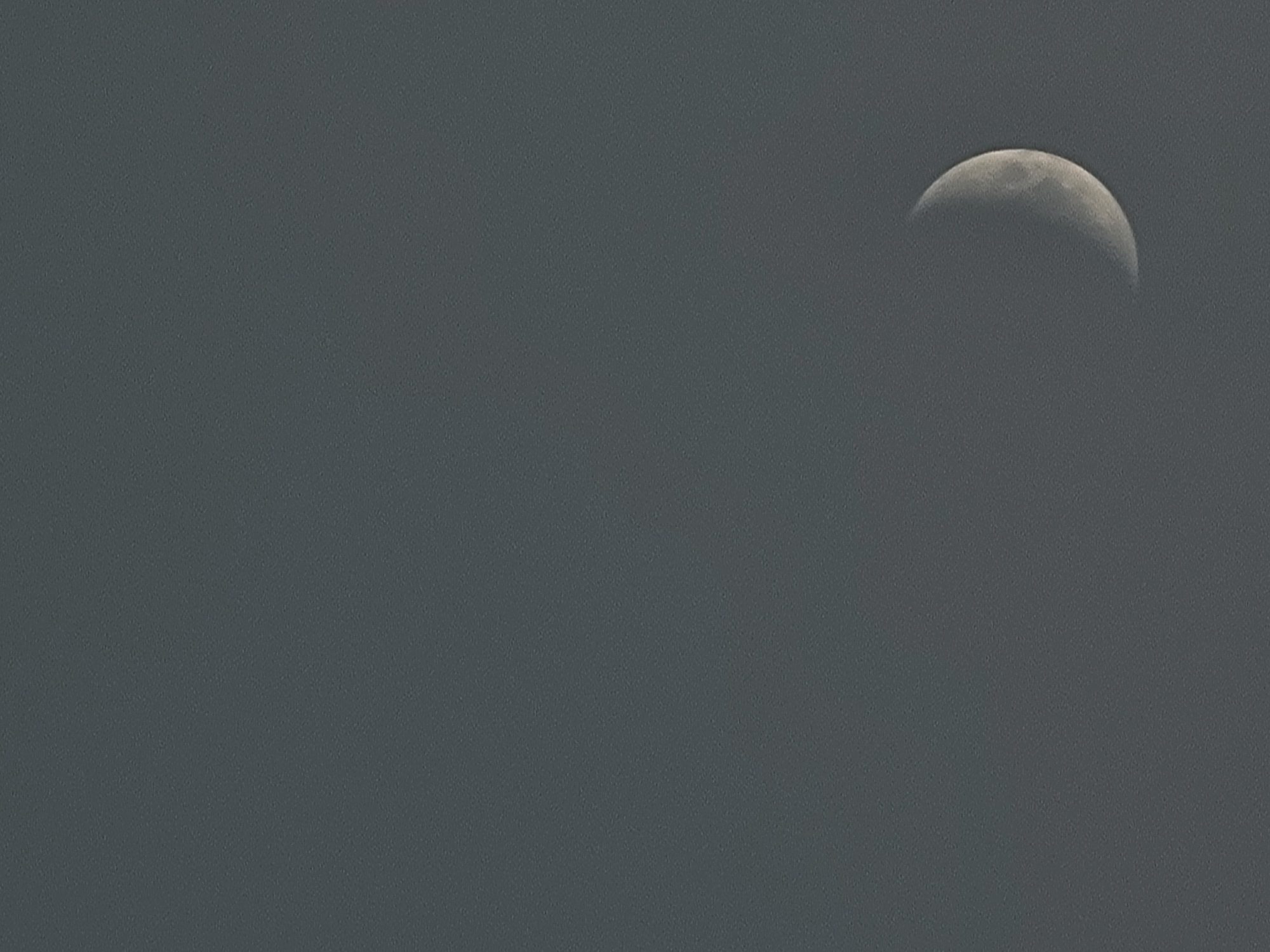 The image is a photograph of a crescent moon in a gray sky. The moon is located in the lower right quadrant of the frame. The sky is overcast and appears to be dusk or dawn. There are no other objects or subjects visible in the image. The location is not specified in the image.