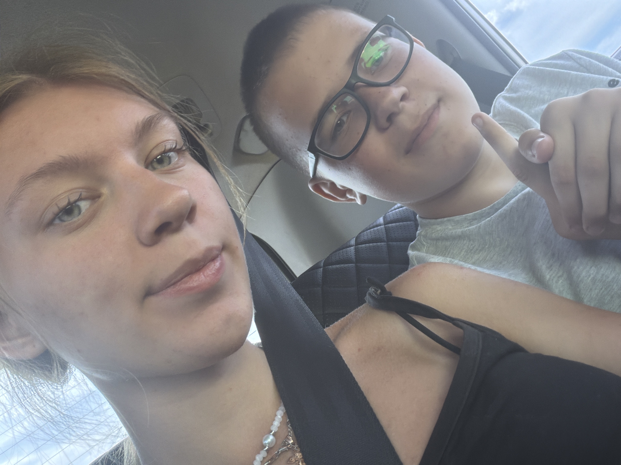 This is a selfie taken inside a vehicle, with two individuals visible. The person in the foreground on the left is a young woman with blonde hair, her face angled towards the camera. She is wearing a black tank top and a necklace. A dark seatbelt crosses her chest. To her right and slightly lower in the frame is a young boy wearing glasses and a light gray t-shirt. He is pointing his index finger towards the camera. The background suggests the interior of a car, with a quilted headrest visible behind the boy. The lighting indicates it is daytime, with sunlight filtering in from outside. There is no information in the image to suggest a location in the city of Uziukovo, Russia.