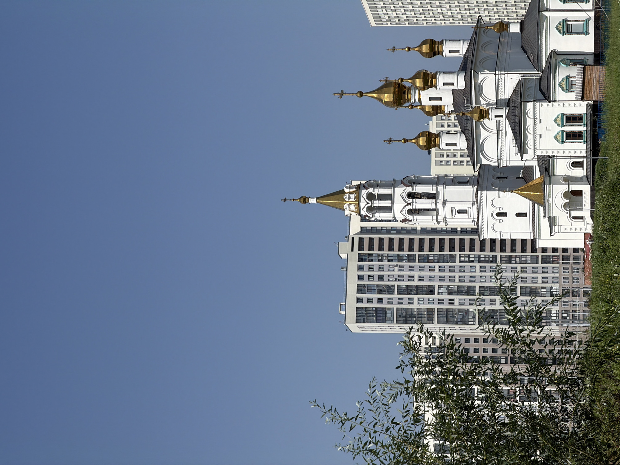 This is a medium shot, outdoor photograph taken on a sunny day, depicting the Church of the Assumption of the Holy Virgin in Yekaterinburg, Russia. The church, with its white walls and golden domes, is situated in the foreground, partially obscured by lush green foliage in the lower left corner. Behind the church, several modern, tall residential buildings are visible, their facades characterized by numerous windows and a neutral color palette. The sky above is a clear, deep blue, devoid of clouds. The image captures a blend of traditional religious architecture with contemporary urban development.