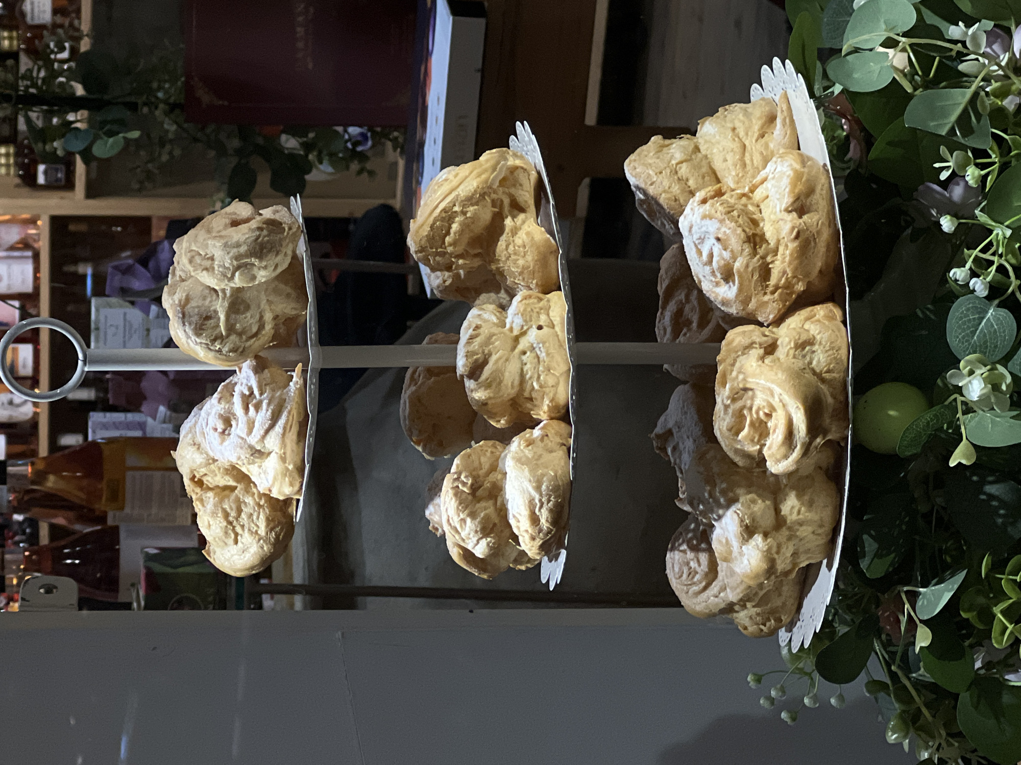 The image shows a three-tiered dessert stand filled with cream puffs, potentially a pastry shop or a dessert display. The stand is metal, with each tier holding multiple cream puffs. Some of the cream puffs are dusted with powdered sugar. The background reveals a shelf stocked with bottles, possibly indicating a store or cafe setting. Greenery is arranged around the dessert stand, adding a decorative touch. The location in Yerevan, Armenia, cannot be definitively determined from the given image.