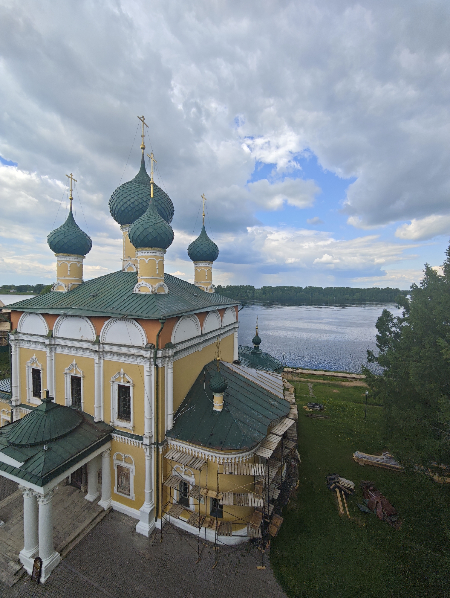 Captured from an elevated perspective, the image presents a detailed view of the Temple of the Assumption in the city of Selivanovo, Russia. The architecture, rendered in yellow and white with green roofing, showcases scaffolding along one side, indicating ongoing renovations. The temple's multiple onion domes, each topped with a cross, dominate the upper portion of the frame. To the right, a body of water stretches toward a treeline in the distance. Debris and construction materials are scattered on the grassy lawn adjacent to the building. The sky is filled with a mix of clouds, transitioning from light blue to a dense, overcast gray.