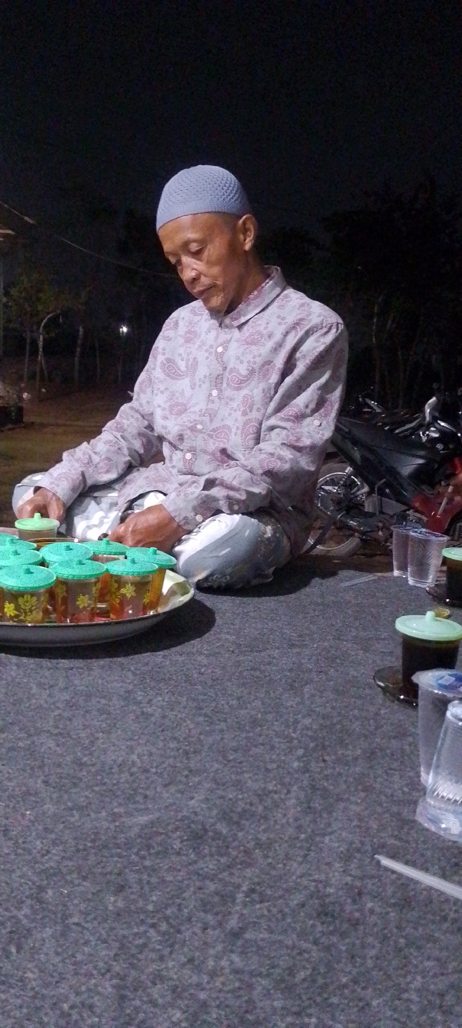 An elderly man wearing a grey kufi and a patterned grey shirt sits cross-legged on a dark grey carpet at night. In front of him is a platter of small glasses with green lids, each containing a liquid. To his right, on the carpet, are several clear plastic cups, some containing dark liquid, and a motorcycle is parked behind him. The background is dark and out of focus, with faint lights suggesting trees and possibly structures. The man appears to be looking down, possibly at the platter of drinks. The scene suggests an informal gathering outdoors at night.