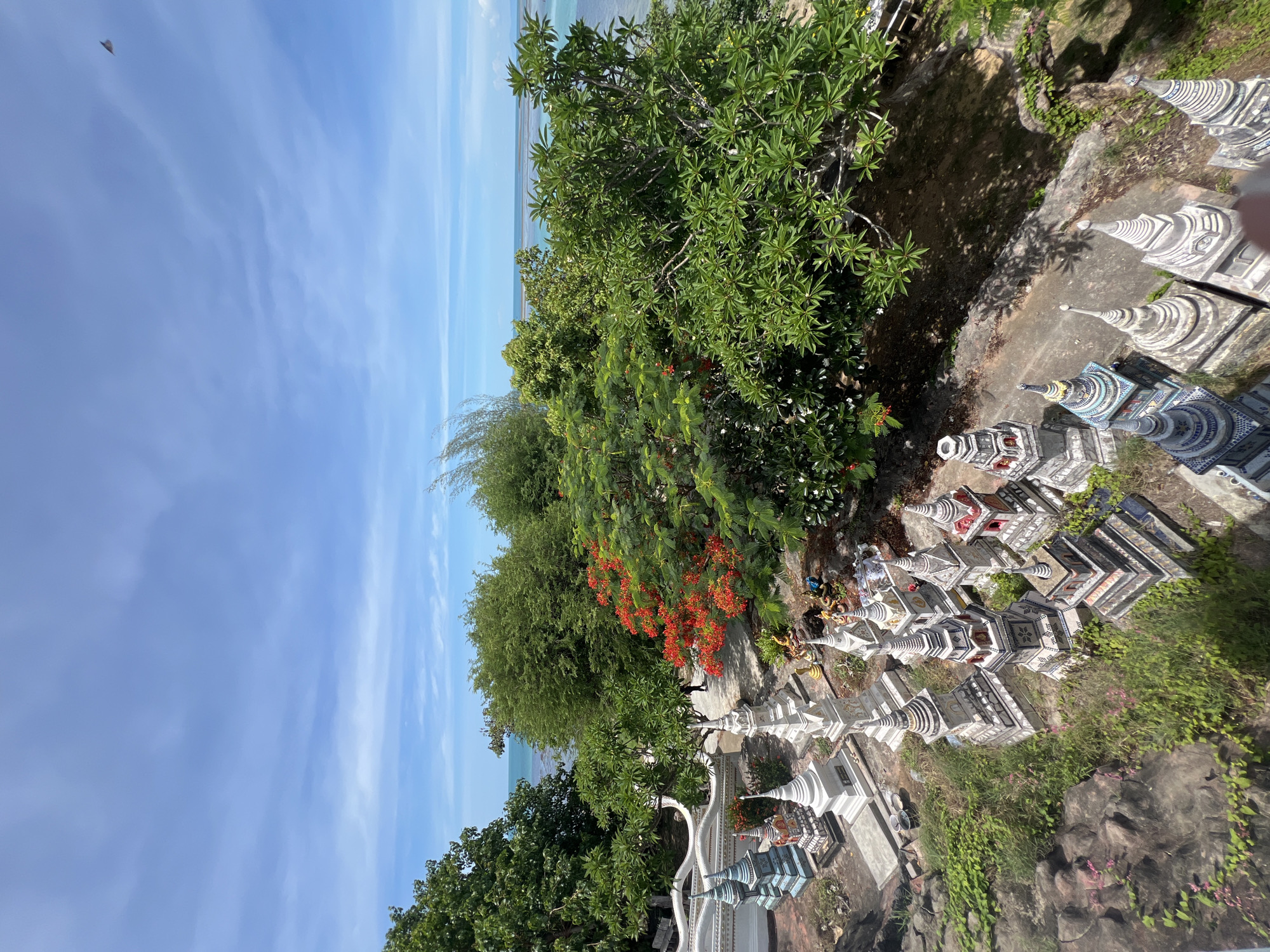 This image shows a view from above, looking down on a collection of small stupas and religious structures, likely in a temple or cemetery. These structures, varying in size and design, are arranged on a sloped area with a stone wall and lush greenery. The background features a clear blue sky with scattered clouds and a glimpse of the turquoise ocean. Trees and vibrant red flowers are visible throughout the scene, adding to the tropical ambiance. The presence of the sea suggests a coastal location, consistent with Baan Hua Thanon, Thailand. The overall atmosphere is one of tranquility and cultural significance.