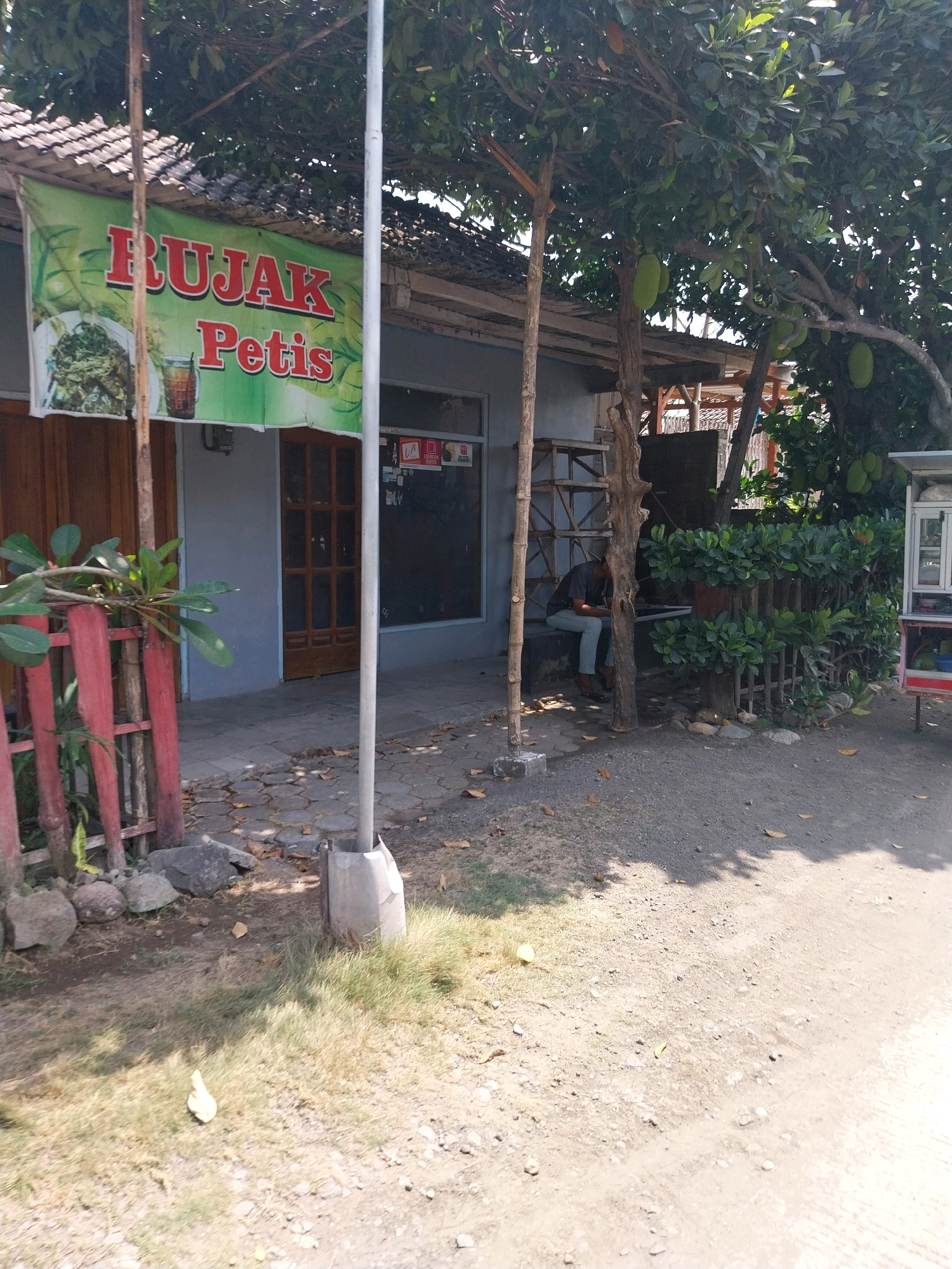 The image shows the exterior of a food stall in Campur, Indonesia, specializing in "Rujak Petis." A green banner with red lettering proclaiming "Rujak Petis" is prominently displayed. To the left of the banner, a plate of mixed vegetables and a glass of iced tea are depicted. The stall itself has a weathered wooden facade and a tiled roof. In front of the stall, a person is seated on a low concrete structure, looking down at a mobile device. A jackfruit tree with several fruits is visible to the right of the stall. The ground is covered with gravel and dry leaves. A red wooden fence runs along the left side of the property, partially obscured by foliage. The overall scene depicts a simple, outdoor food establishment in a tropical setting.