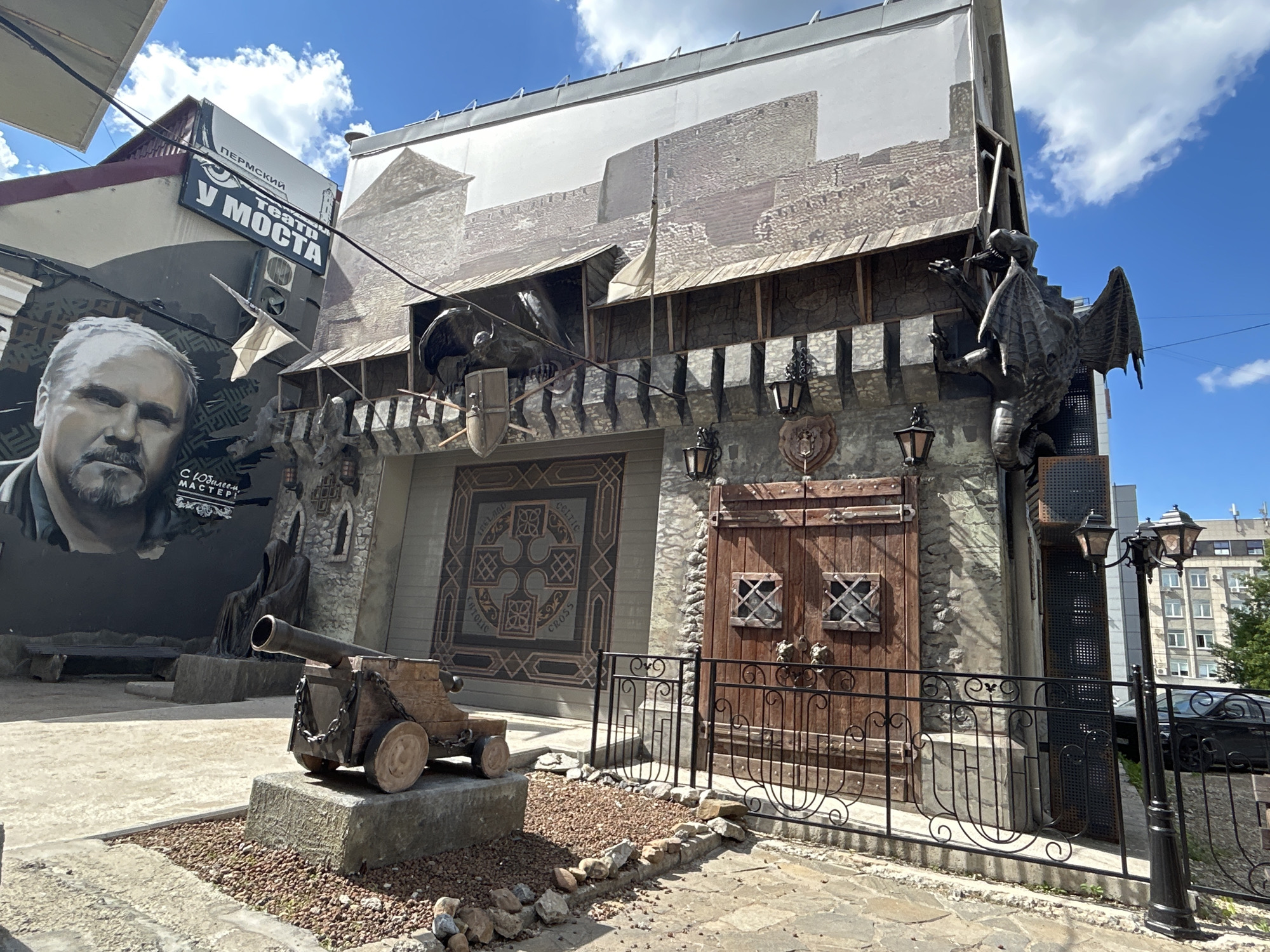 The image shows the facade of a building in Perm, Russia, designed to resemble a medieval castle. A large mural on the left features a portrait of a man with a mustache and beard, alongside text in Russian that appears to be related to a theater. The building itself is adorned with architectural elements that evoke a fortress, including stone textures, wooden doors with iron accents, and decorative gargoyles or dragon sculptures. A miniature cannon is placed in the foreground on a stone base, and a wrought-iron fence runs along the pathway in front of the building. The sky is blue with scattered clouds, and a lamppost with multiple lights stands to the right. In the background, modern buildings are visible, suggesting an urban setting.