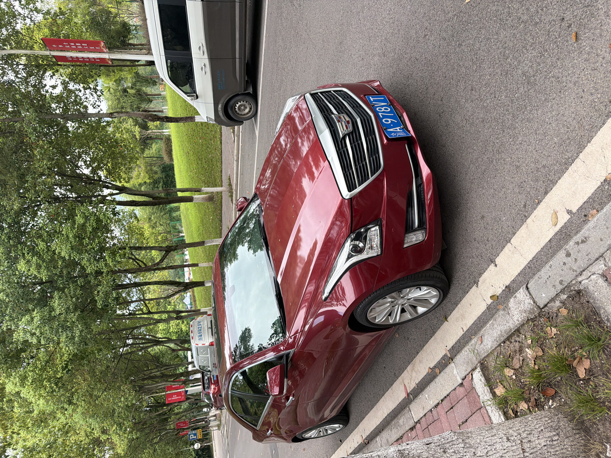 A red Cadillac sedan is parked on the side of a street in Jiangjin, China. The car is facing right, with its front end slightly angled towards the viewer. It has a blue license plate with white characters that read "A-978VT". The street is lined with trees, and there is a grassy slope on the right side of the image. A white and brown van is parked further down the street behind the Cadillac. A red banner with Chinese characters hangs from a pole on the left side of the street. The ground is paved with asphalt, and there is a concrete curb and sidewalk.