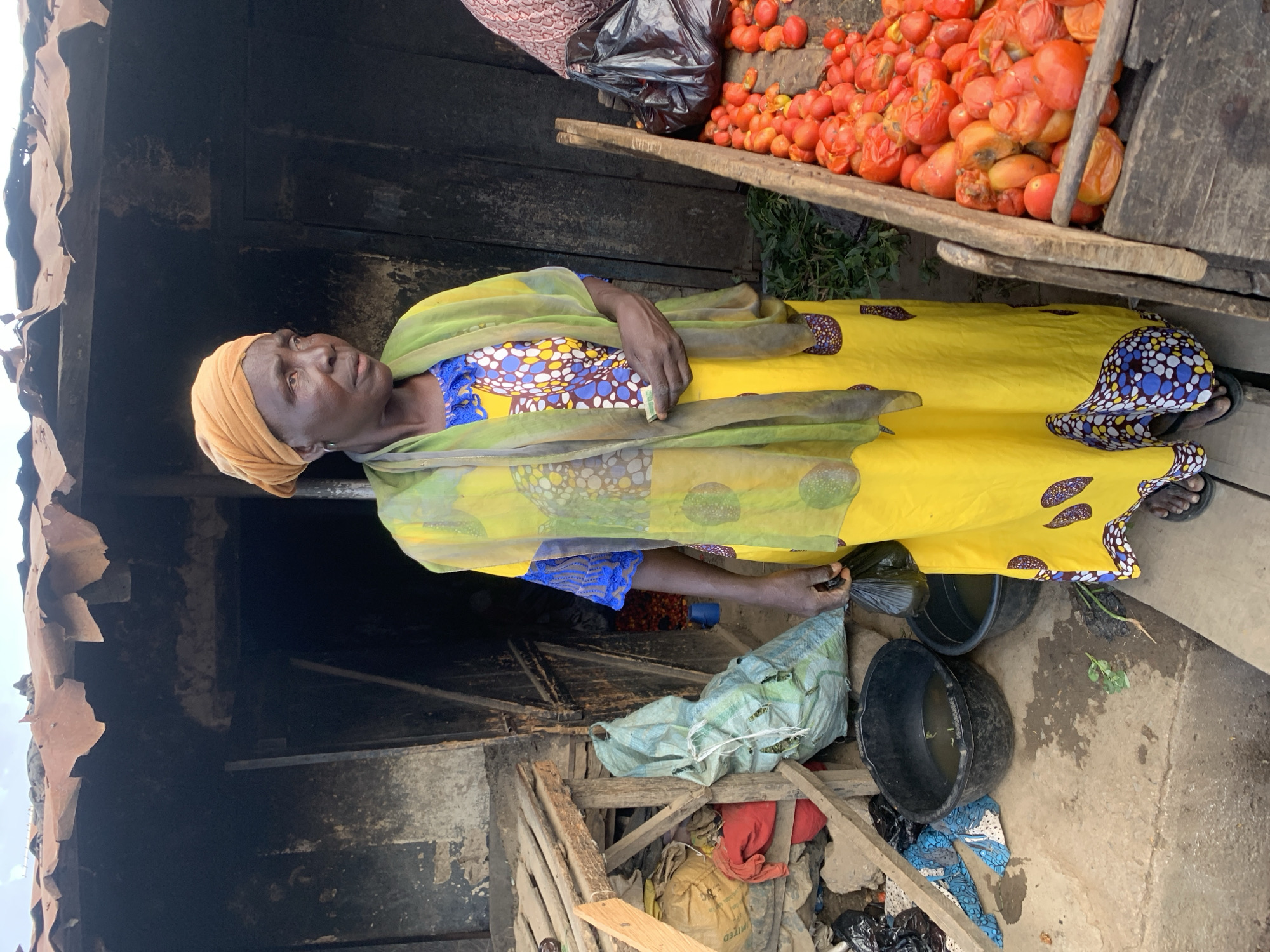 In Jos, Nigeria, an elderly woman stands in a dimly lit outdoor market stall, potentially her place of business. She is dressed in a yellow and patterned dress, with a light green shawl or scarf. Her head is wrapped in a light brown or beige head covering. To her right are several piles of red tomatoes, some of which appear bruised or damaged. In front of her are several black plastic buckets. Below her and to her left is a rough wooden structure with other discarded items. The background behind the stall is dark, and the roofing of the stall is made of corrugated metal.