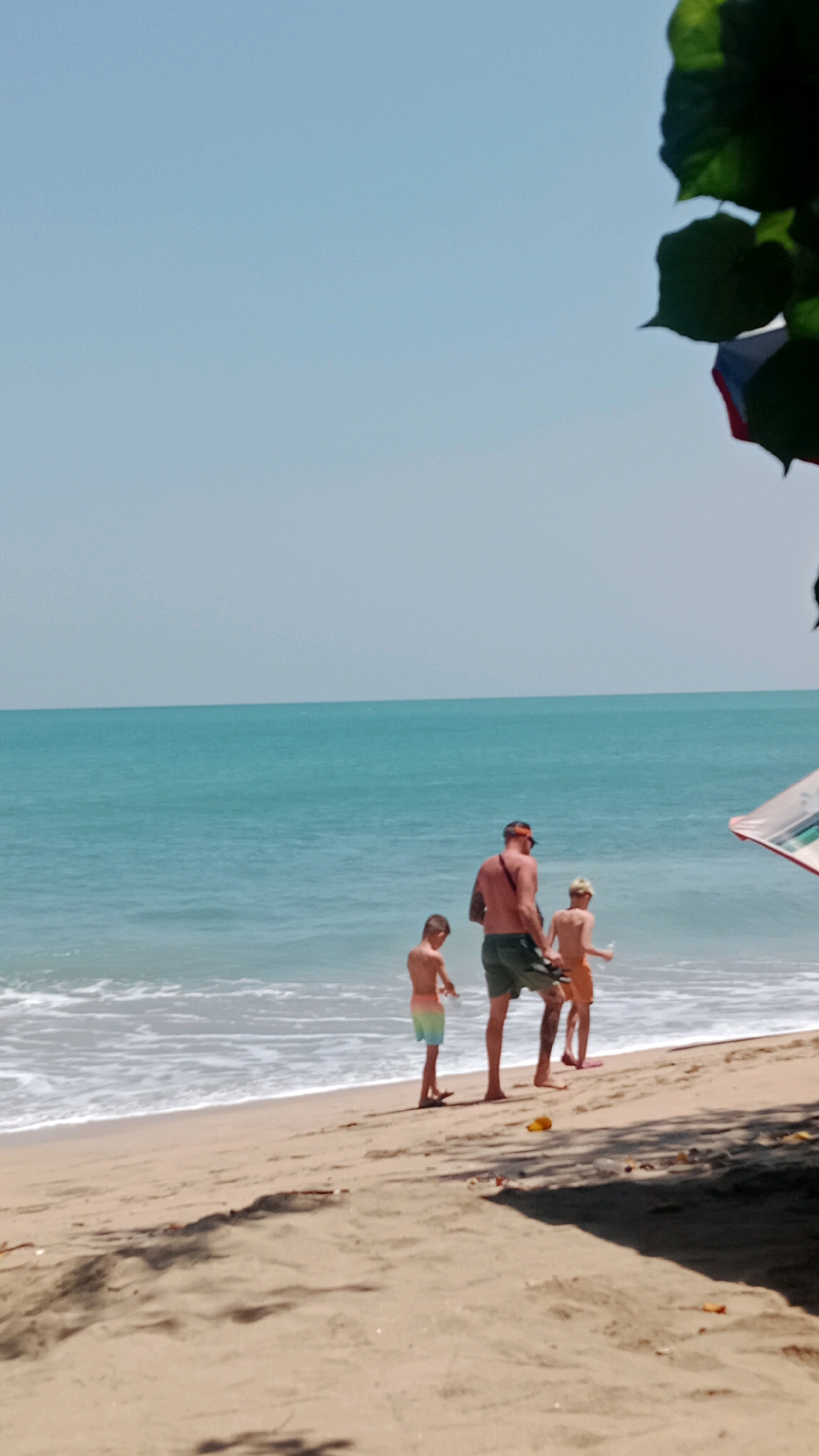 In Kuta, Indonesia, on a sandy beach, a man and two boys are standing near the ocean. The man, facing away from the camera, wears a reddish-orange hat and green shorts, with a strap across his chest. The boy to his left, also facing away, is wearing multicolored swim trunks. The boy to his right, also facing away, is wearing light-colored shorts and a yellow cap. The ocean, with gentle waves, stretches to the horizon under a clear sky. A portion of a beach umbrella is visible on the right side of the frame. The sand in the foreground shows shadows and scattered debris.