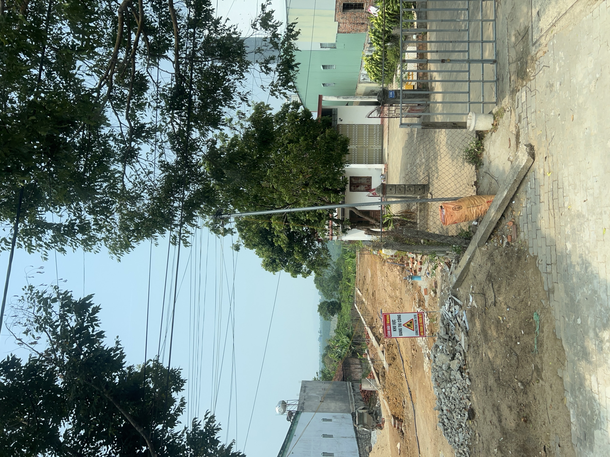 The image depicts a construction site in Đăk Hà, Vietnam. A warning sign reads "CONSTRUCTION SITE. Safety comes first." The ground is torn up with piles of rubble and dirt. In the background, there is a row of buildings, including houses and a taller structure. Trees flank both sides of the view, and power lines stretch across the sky. A small white bucket sits just beyond the fence. A small brown bag sits next to the fence.
