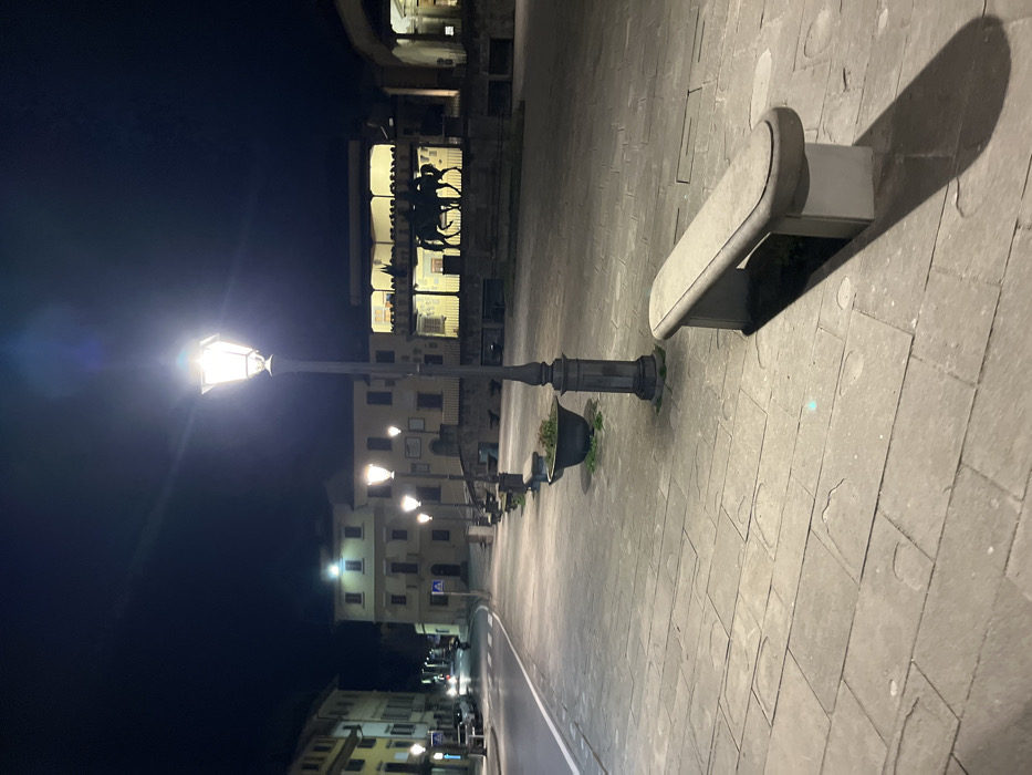 The image captures a quiet, illuminated street or plaza in Fiesole, Italy, at night. The perspective looks down a paved area towards a street lined with historic-looking buildings.
In the foreground on the right, a smooth, light-colored stone bench casts a long, distinct shadow onto the patterned stone paving. To the left of the bench, a traditional black lamppost with an ornate base and a planter containing sparse greenery emits a bright white light, causing a lens flare effect. The lamppost also casts a noticeable shadow. A series of similar lampposts extends into the background, providing consistent street lighting.
On the left side of the frame, a road or thoroughfare is visible, bordered by more buildings. Across this road, multi-story buildings with various illuminated windows can be seen. A prominent building in the mid-background features an architectural structure, possibly a loggia or balcony, on which a dark silhouette resembling a statue of an equestrian figure or a chariot is visible. The sky is dark, confirming the nighttime setting. The ground is paved with light-colored, rectangular stone slabs. The scene is devoid of people, suggesting a late or early hour, and conveys a serene atmosphere. No legible text is visible.