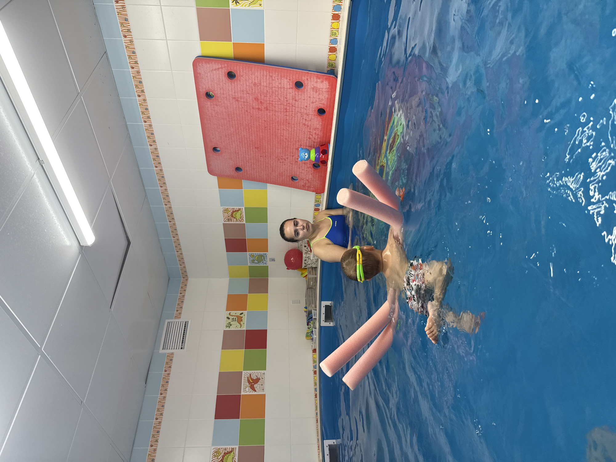 The image shows two people in a swimming pool. One person, an adult woman wearing a blue swimsuit, is partially submerged in the water and looking at the camera. The other person is a child, wearing swim trunks and goggles, swimming in the pool. The child is holding onto two pink pool noodles. The pool area is decorated with colorful tiles featuring images of animals and geometric patterns. A large red flotation device with holes is visible near the edge of the pool, along with a small toy. The lighting suggests it is daytime, and the indoor setting of a swimming pool is evident. The overall mood appears to be one of recreation or swimming lessons. There is no visible text in the image.