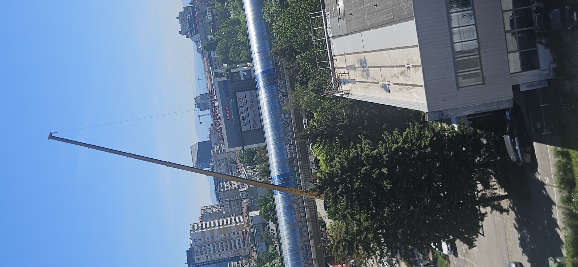 The image shows an aerial view of a cityscape in Sofia, Bulgaria, under a clear blue sky. A construction crane extends across the frame from left to center. Adjacent to the crane's base is a large, enclosed pedestrian bridge with blue and silver exterior panels. The bridge runs along a commercial area. In the background, there's a building with a prominent sign that reads "ДОВЕРИЕ," which appears to be a brand name. The commercial area transitions into a dense residential area, with numerous multi-story buildings visible. A commercial building with what appears to be an "easybox" sign is visible to the right. Trees surround the base of the building and cars are parked in the parking lot. The image suggests ongoing construction or urban development within the city.