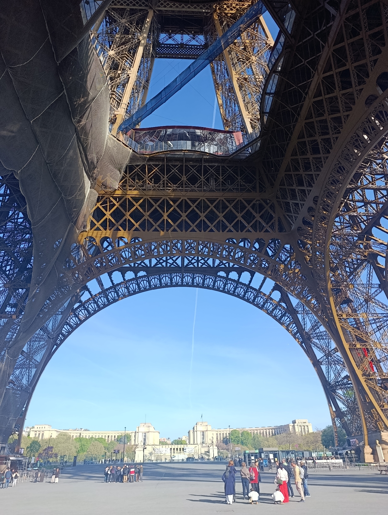 The image shows the view looking up from underneath the Eiffel Tower in Paris, France. The tower's metal framework arches overhead, partly covered by netting on one side. Above the arch, the structure continues upward, with a visible observation deck enclosed in glass. The sky is blue. At ground level, a paved area is populated by a small group of people standing around. In the background, buildings can be seen across a plaza or park.