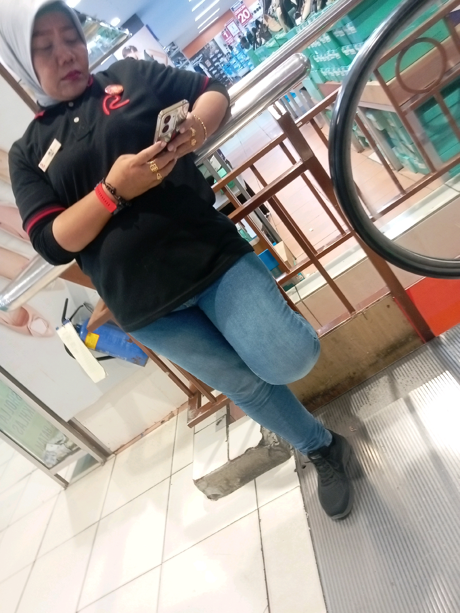 A woman wearing a black polo shirt with red trim, blue jeans, and a grey hijab is standing on an escalator in a shopping mall. She is holding a smartphone in her right hand and looking at it. She has a red wristband on her left wrist and several gold rings on her fingers. The escalator appears to be in a public area, possibly a department store, with displays of merchandise visible in the background. The scene is indoors with bright overhead lighting. The location is identified as Batam, Indonesia.