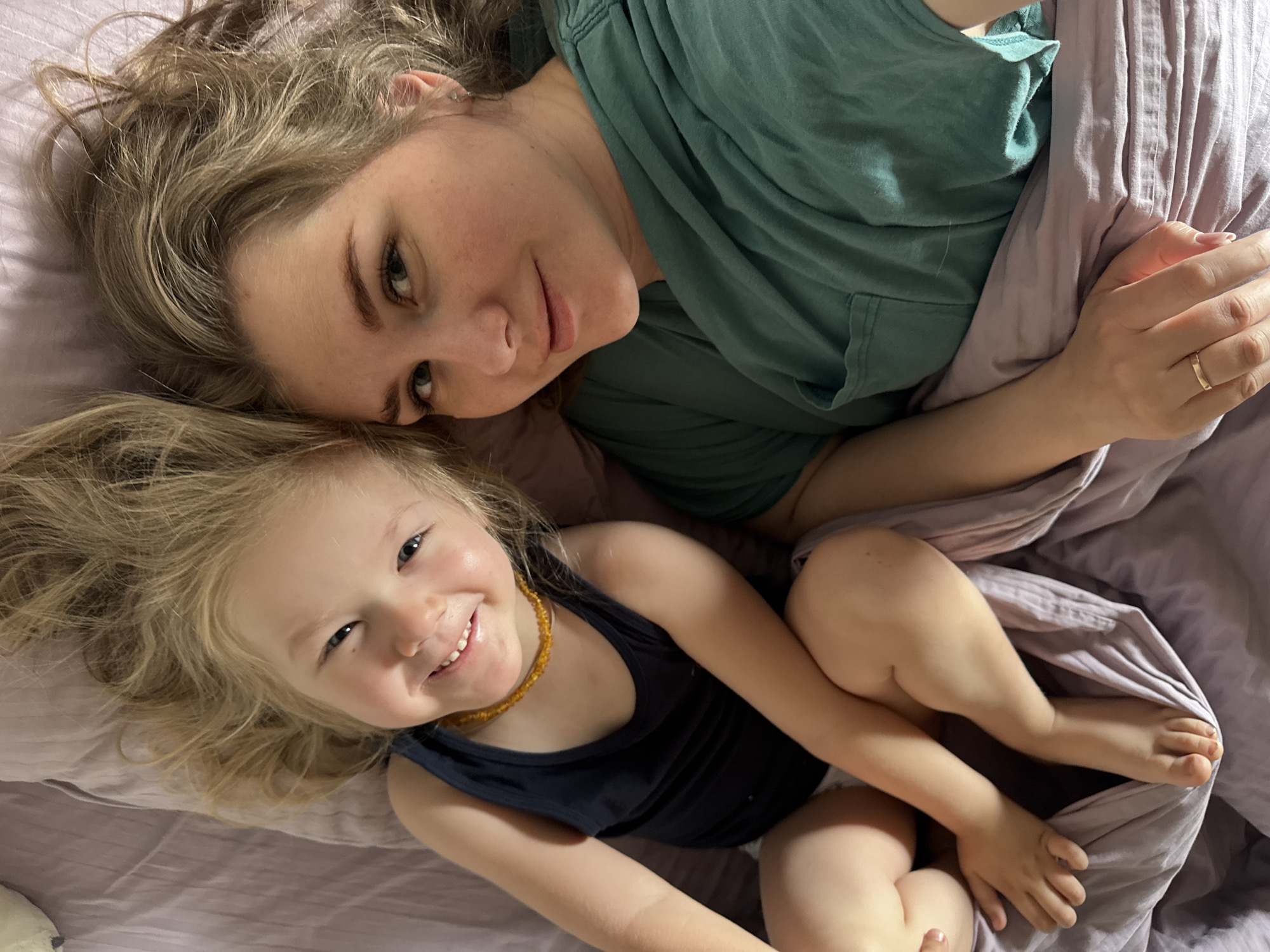 The image depicts a blonde woman and a young blonde child lying on a bed covered with lavender-colored bedding. The woman wears a green t-shirt and a wedding ring on her left ring finger. The child wears a dark blue tank top and a brown amber necklace. Both are smiling and looking at the camera. The location is not identifiable as being in the city of Ферма, Russia.
