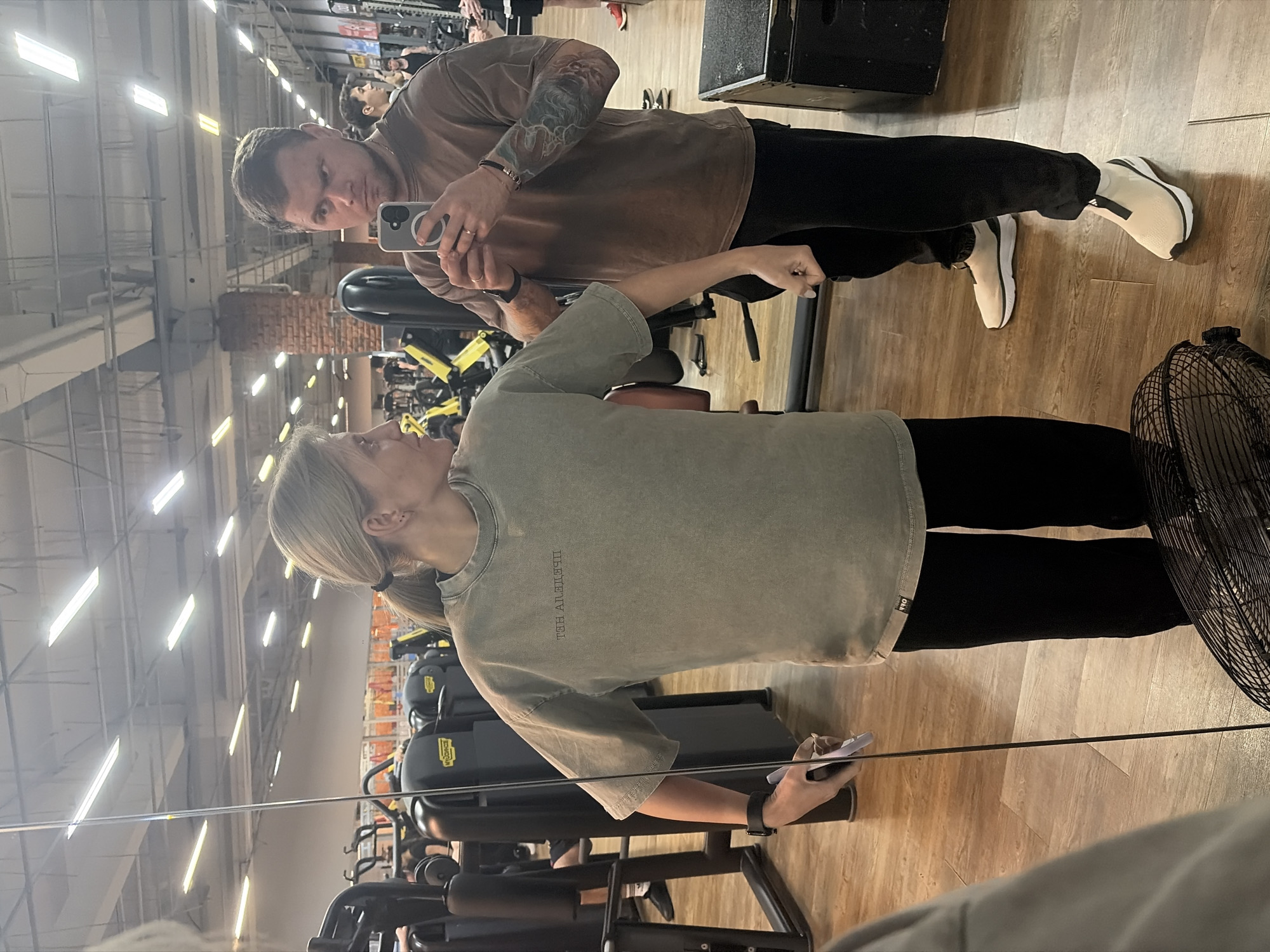 A man and a woman are posing for a picture in a gym. The man, on the left, is holding a phone up to take the picture. He is wearing a brown t-shirt, black pants, and white shoes. He has tattoos on his arms. The woman is standing next to him, wearing a green t-shirt and black pants. In the background, there are exercise machines and other people working out. A large fan is partially visible on the right. The floor is wooden. The location is a gym in the city of Chaikovsky, Russia.