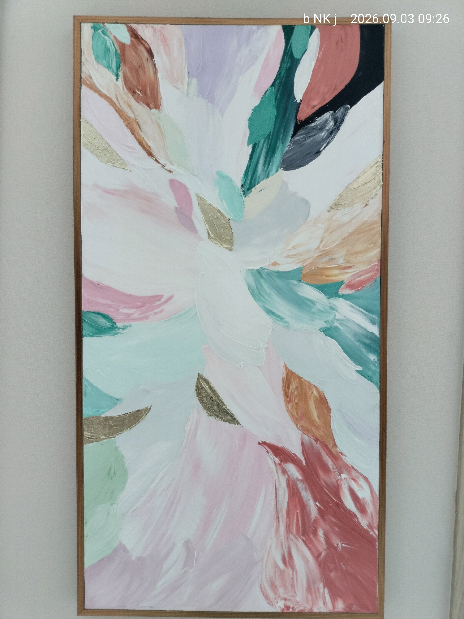A vertical, close-up shot shows a framed abstract painting with thick brushstrokes. The artwork features a central burst of white, surrounded by soft pastel colors like pink, lavender, mint green, and light brown, interspersed with metallic gold leaf accents. The painting is framed in a simple, light brown wooden frame and is hung against a plain, light grey wall. The overall impression is one of light, airy movement. The location is in South Jakarta, Indonesia.