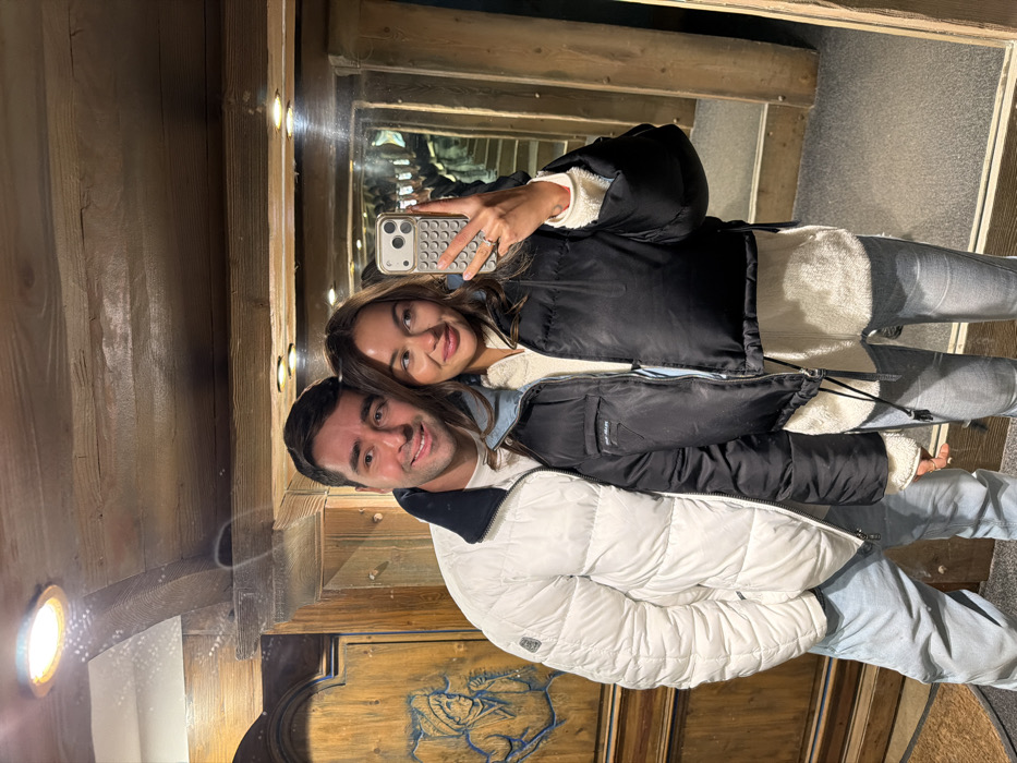A couple takes a selfie in a room with wood paneling. The man wears a white puffer jacket, and the woman is in a black puffer jacket and holds a phone. A hallway is visible in a mirror behind them. The woman holds up a peace sign while taking the selfie.