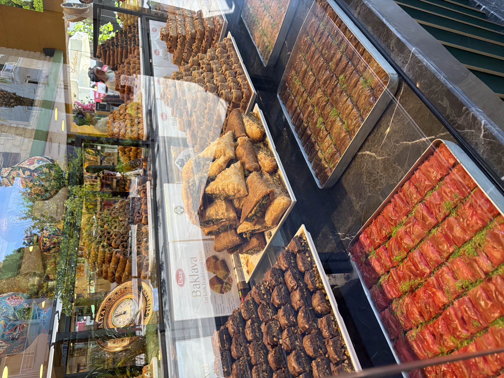 The image shows a display window of a baklava shop, featuring a variety of baklava pastries. The pastries are arranged in trays and on tiered stands, with different textures and toppings visible. Some baklava appears to be covered in chocolate or nuts, while others are sprinkled with green pistachios. Behind the baklava, there are decorative vases and a clock. Reflections on the glass reveal the exterior environment, including trees, buildings, and people walking by. The overall scene suggests a brightly lit shop with a diverse selection of sweet treats.