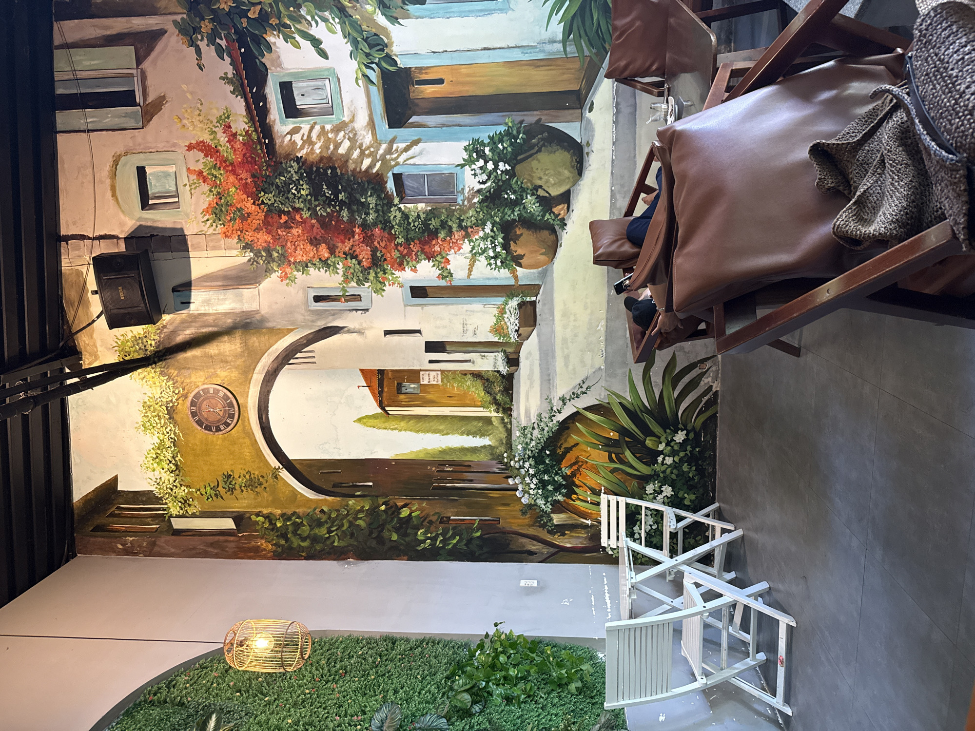 The image depicts the interior of a cafe with a large mural on the wall, creating an illusion of an outdoor scene. The mural features an arched pathway with buildings, plants, and a clock. In the foreground, there are two white folding chairs and a table set on a dark tiled floor. To the right, there are brown leather lounge chairs with a person relaxing in one, holding a phone. A woven bag rests on another chair. The cafe is decorated with a vertical garden wall on the left and a hanging light fixture. A speaker is mounted on the mural. The overall atmosphere suggests a relaxed, indoor seating area designed to evoke an outdoor European setting. The cafe is likely located in Nha Trang, Vietnam, given the provided context.