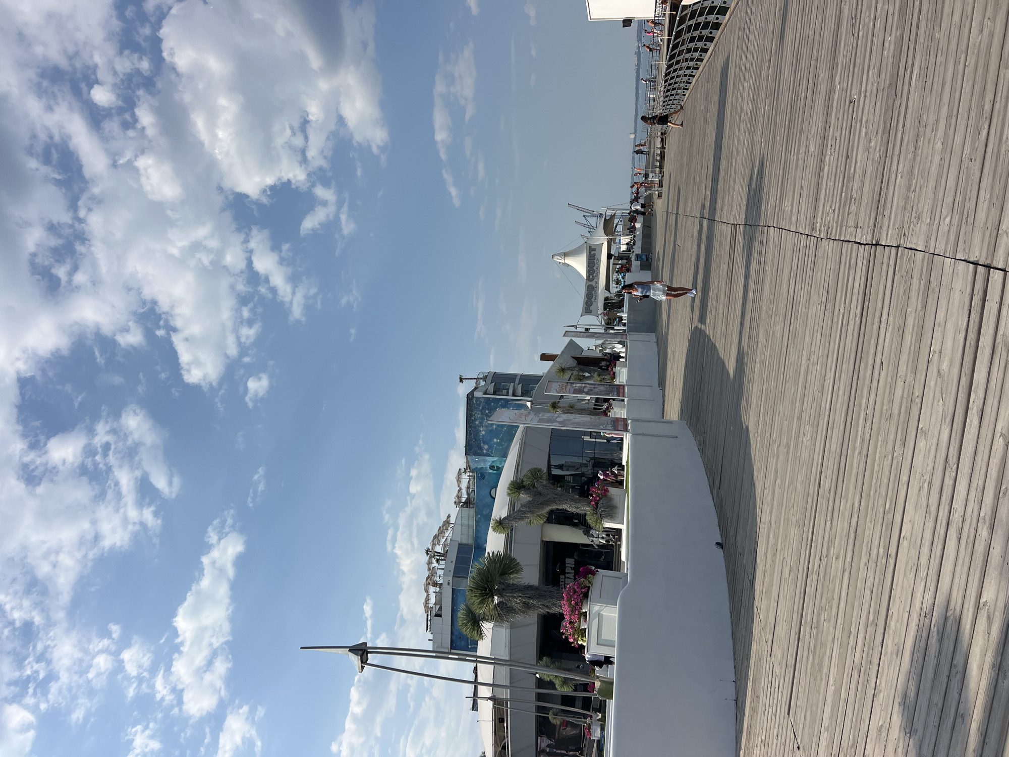 The image depicts a boardwalk along the Black Sea coast in Odesa, Ukraine. The boardwalk is constructed of weathered wooden planks and curves to the right, leading towards a distant structure. To the left of the boardwalk, a modern, white-walled building with large windows is visible, featuring palm trees and flower arrangements in front. This building appears to be a commercial establishment, possibly a restaurant or club, with signage in Cyrillic. Further down the boardwalk, a white tent-like structure with the words "FOOD COURT" is present, indicating a dining area. Several people are walking along the boardwalk, dressed in casual summer attire. The sky is blue with scattered white clouds, suggesting a sunny day. The sea is visible in the distance to the right, with a faint outline of a ship. The scene conveys a lively atmosphere of a popular seaside resort or promenade.