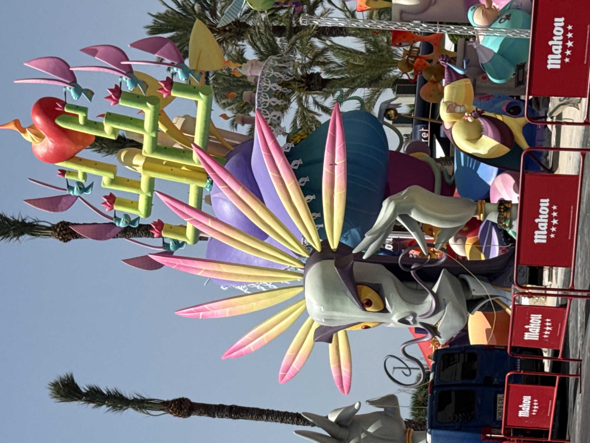 An outdoor scene with a large, brightly colored sculpture of a fantastical creature, possibly a dragon or a monster, with a skull-like head adorned with feathered headwear. The sculpture is very elaborate and features multiple segmented arms and colorful elements. In the background, palm trees and a clear blue sky are visible. In the foreground, there are red barriers with white text that reads "Mahou" and stars. A blue van is parked behind the sculpture, and other parts of the sculpture, including figures and abstract shapes, are visible. The overall impression is of a festive or celebratory display. The location is Unknown, Unknown.