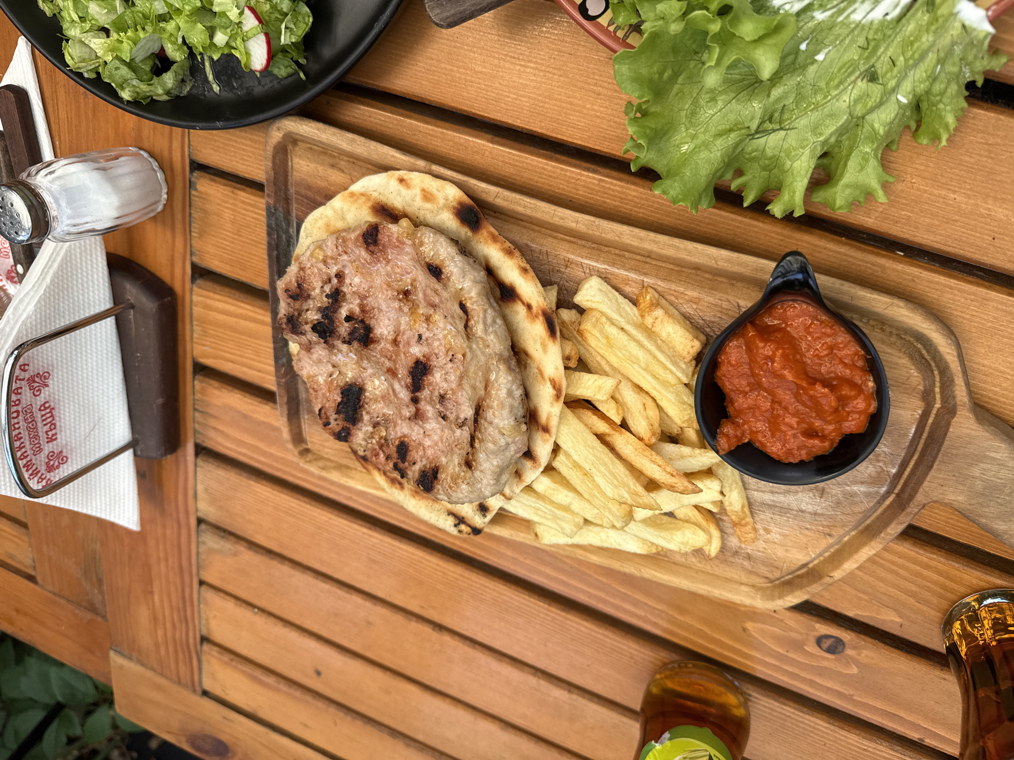 A top-down shot shows a meal on a wooden table at a restaurant in Sofia, Bulgaria. The meal includes a grilled patty on flatbread, french fries, a small dish of red sauce, and a salad. There are also two bottles of amber liquid, likely beer, and a salt shaker. The restaurant's name, written in Cyrillic, is visible on a napkin holder.