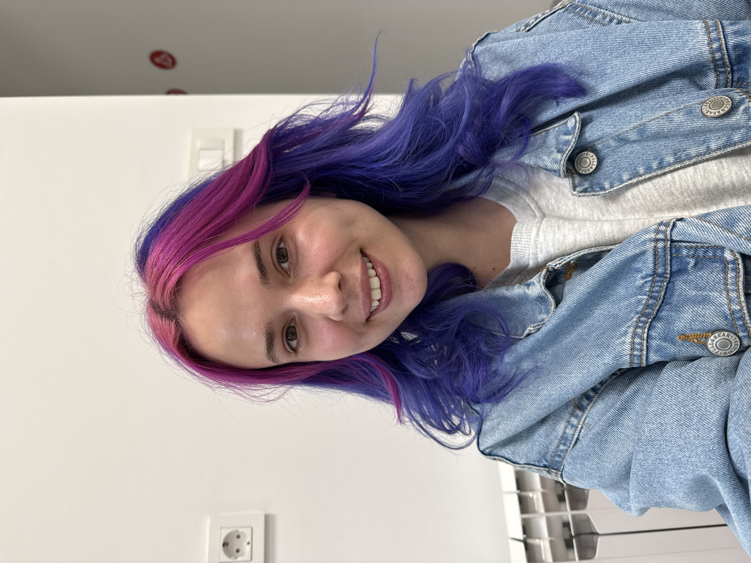 A young woman is featured in a close-up, waist-up portrait, rotated 90 degrees counter-clockwise. She has striking two-tone hair, with vibrant magenta-pink framing her face and at the roots, transitioning to a bright purple-blue through the lengths. Her hair appears voluminous and slightly windswept. She looks directly at the camera with a wide, cheerful smile, revealing her teeth. Her skin is clear, and her eyes are dark.
She is dressed in a light grey crew-neck top, layered under a classic light blue denim jacket. The jacket has visible seams, stitching, and metal buttons, some of which are clearly embossed with the text "BEAR JEANS" and "BEAR."
The background consists of plain white walls, indicating an indoor setting. On the upper left wall, there is a white light switch or cover. A white European-style electrical outlet with two circular holes is visible on the lower left wall. A small red circular object is barely discernible high on the left wall/ceiling. Part of what appears to be an air conditioning unit or vent is visible in the lower right corner. The lighting is bright and even. The scene captures a friendly moment, likely a selfie, occurring in the city of Zagreb, Croatia.